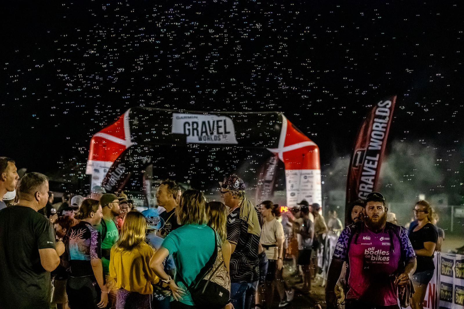 A crowd gathers at night under a lit "Gravel" archway. Participants wearing cycling gear are visible, with some people looking towards the event. Bubbles float in the air, adding to the festive atmosphere.