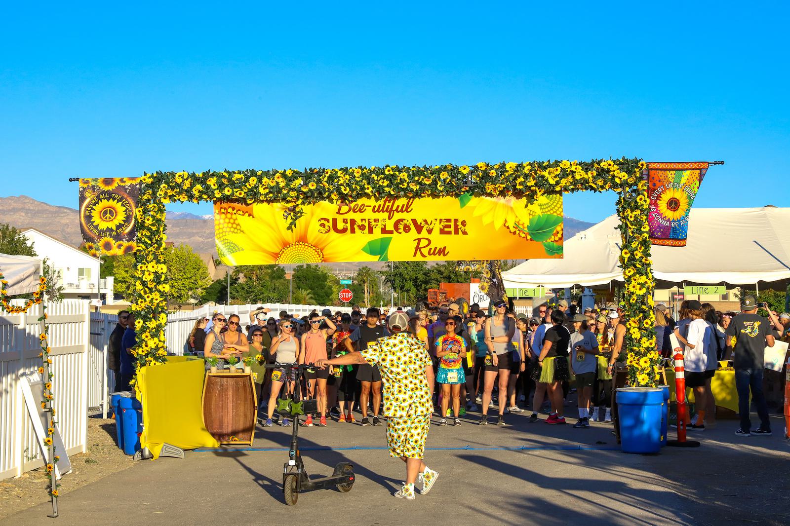 Participants gather under a floral arch at the Sunflower Run event. The arch features yellow sunflowers and a bright sign. Tent booths and staff are visible, with clear blue sky in the background. One person walks with a megaphone.