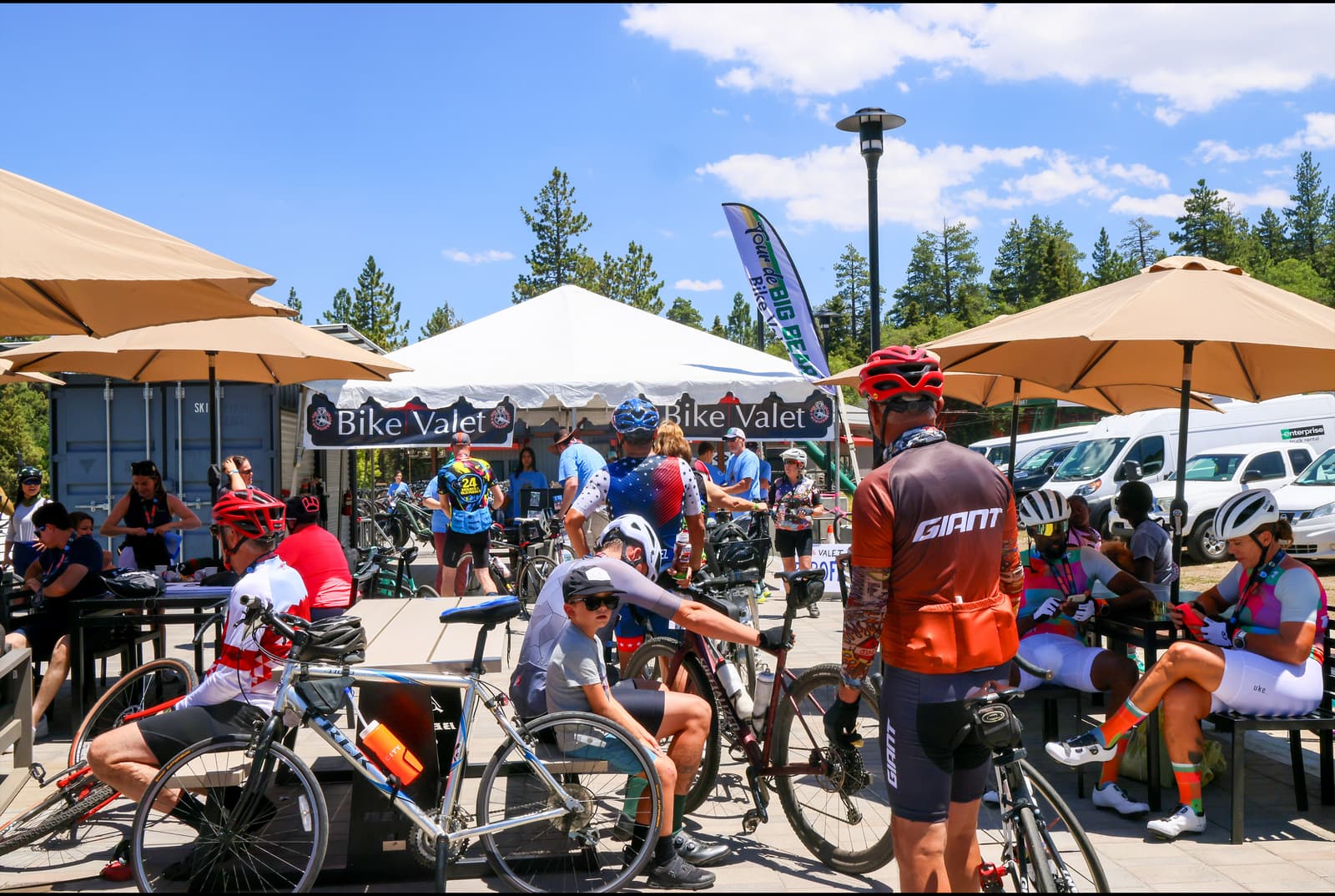 Cyclists relax at a bike valet station outdoors. They're under umbrellas, with some seated and others standing beside their bikes. A banner reads "Bike Valet," and trees are visible in the background. Bright, sunny weather enhances the scene.