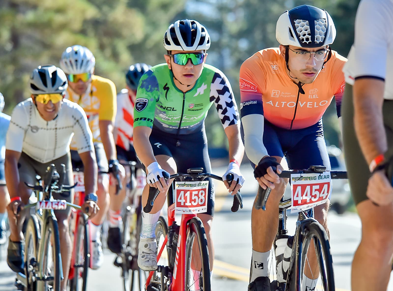 Cyclists competing in a race on a sunny day. The focus is on two leading cyclists wearing helmets and glasses, one in a green jersey and the other in an orange jersey. More cyclists are visible in the background, with trees lining the road.