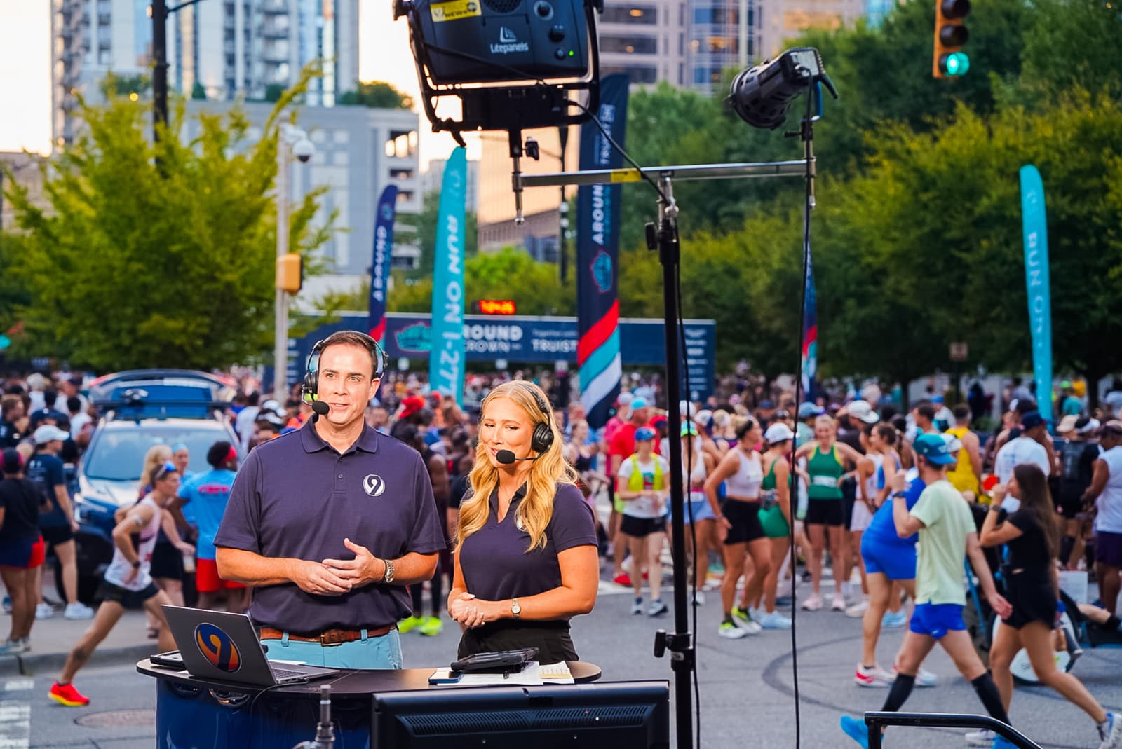 Two broadcasters wearing navy shirts stand at a desk with microphones, reporting live from a bustling outdoor marathon event. Behind them, a crowd of runners prepares to start. Bright, sunny day with trees and tall buildings in the background.