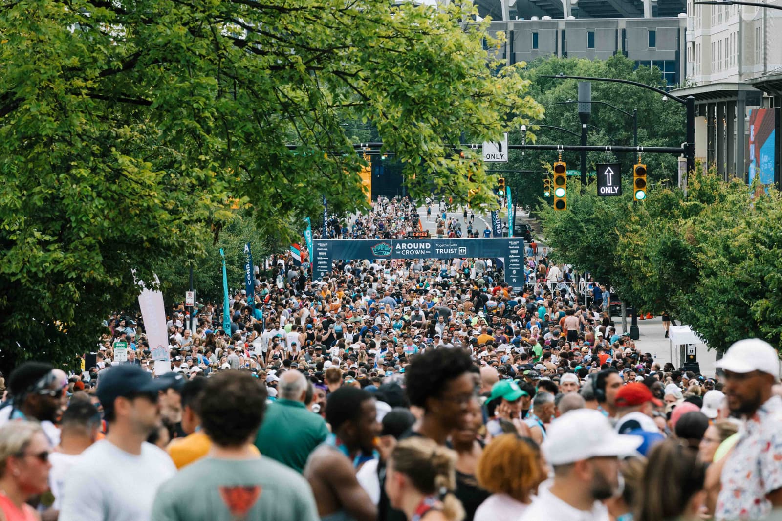 A large crowd gathers on a city street for a marathon event. A banner reading "Around" is visible in the background. Trees line the street, and traffic lights show green and yellow. People wear casual and athletic attire.