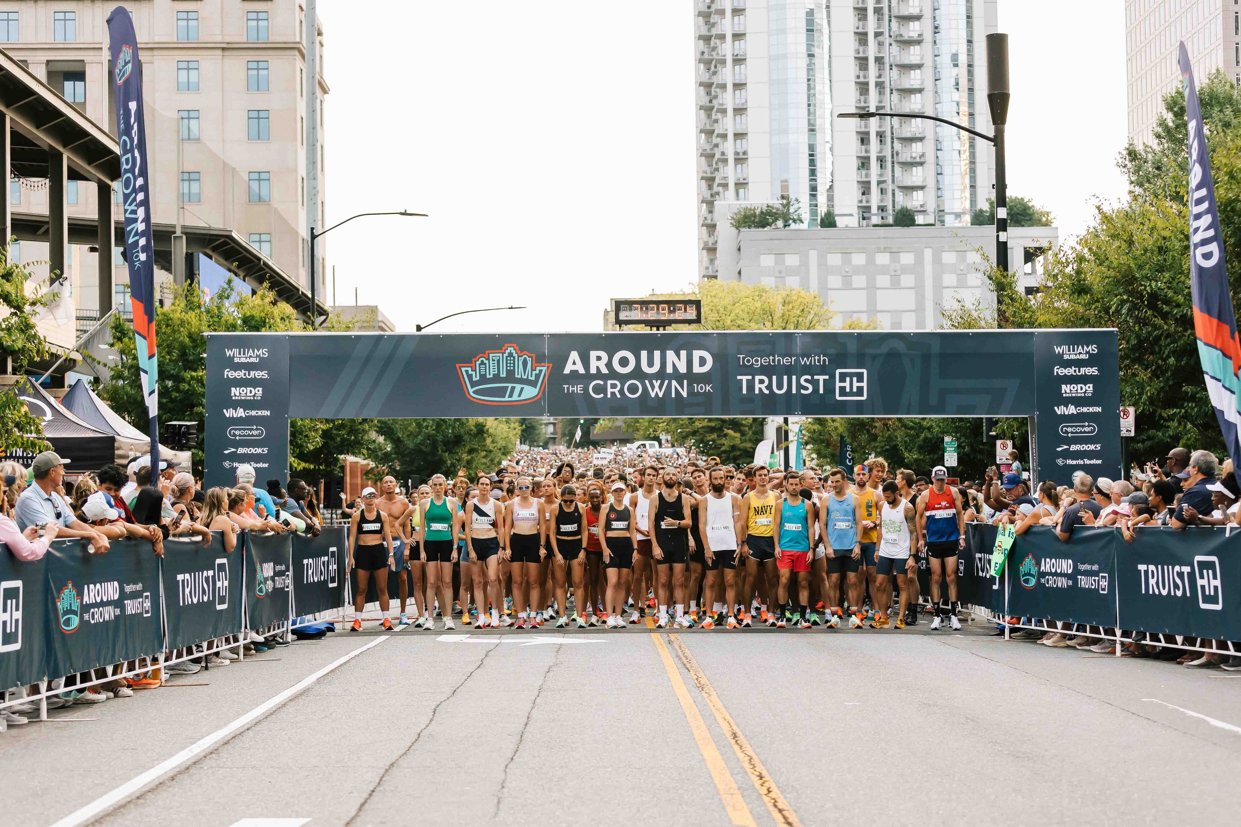 Participants gather at the starting line of a marathon event titled "Around the Crown 10K" in an urban setting, with tall buildings and trees in the background. Banners and barriers display sponsor logos, and spectators line the street.