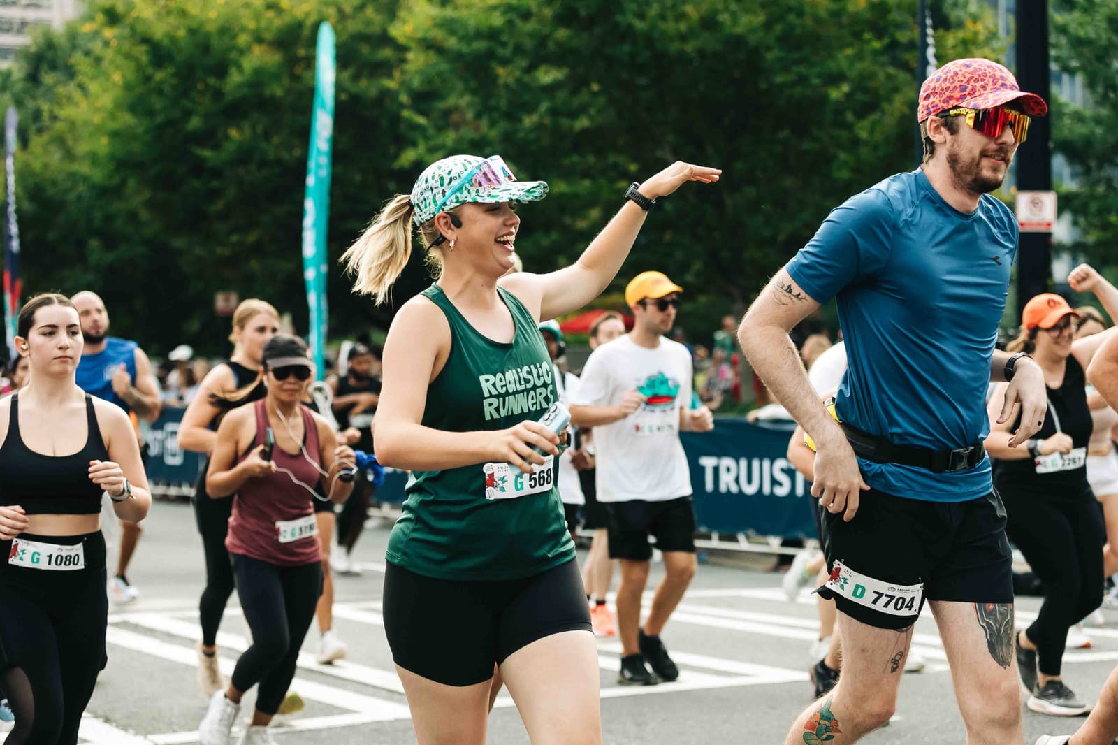 Runners participating in a race on a city street. A woman in a green tank top and patterned cap smiles while running, surrounded by others in athletic wear. Trees and a crowd line the route, with a race banner visible in the background.