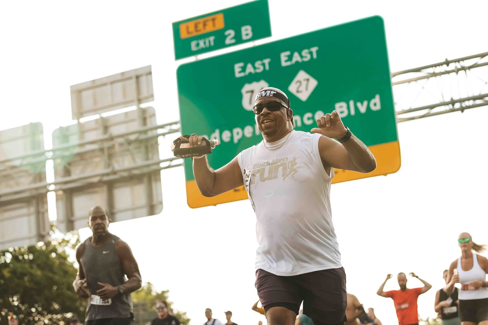 A man in a white tank top and sunglasses smiles while running, holding a smartphone. Other runners are in the background. A large green road sign indicating "Exit 2B" and directions to "East Independence Blvd" is overhead.
