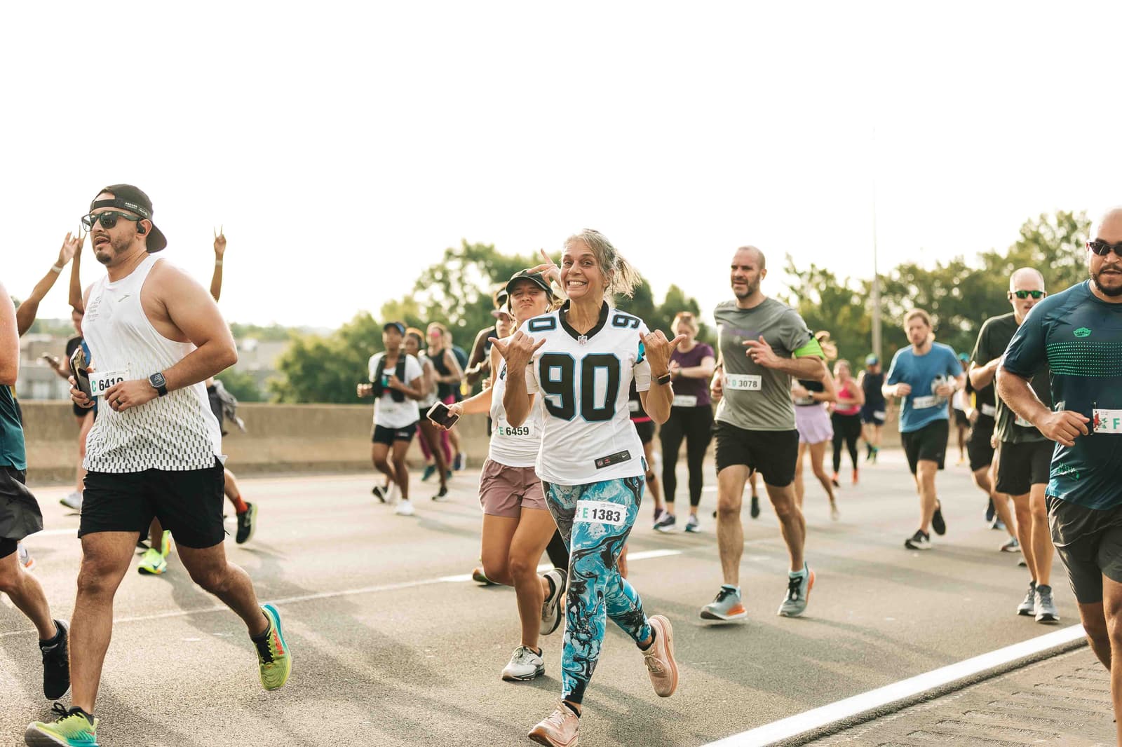 A group of runners in athletic gear participate in a marathon race on a sunny day. A woman in the foreground wearing a "90" jersey smiles and waves at the camera. Other runners are visible in the background on the road.