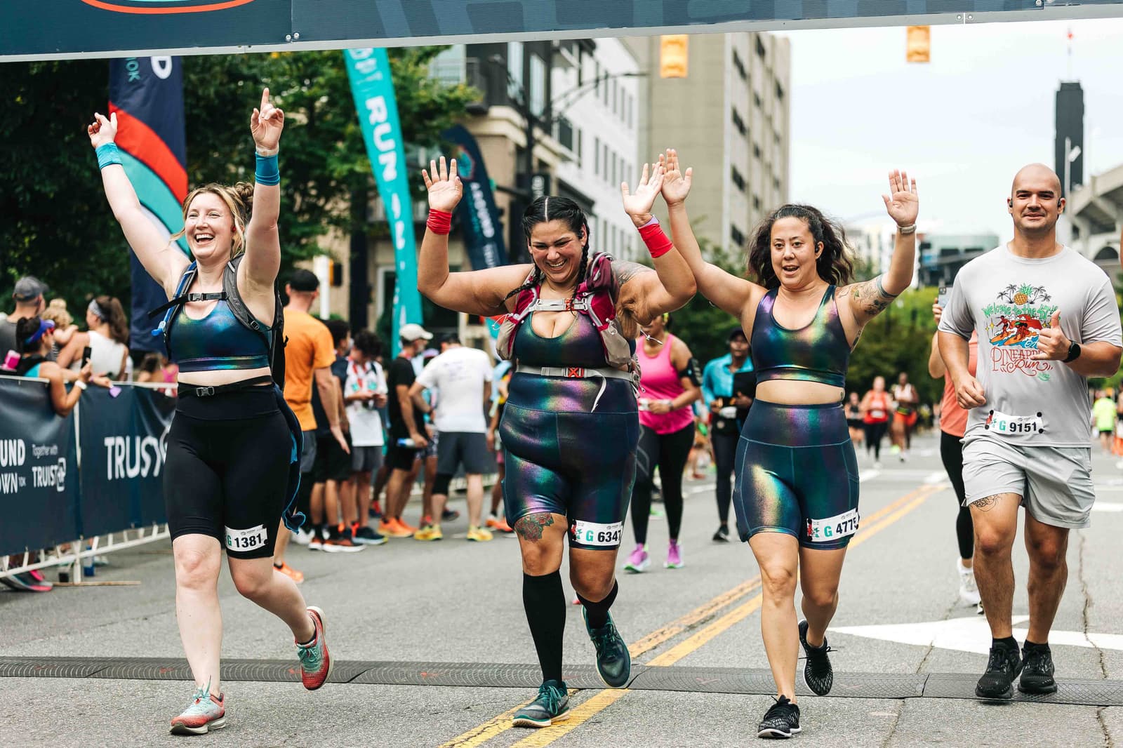 Four people in colorful athletic outfits celebrate as they near a race finish line. They hold their arms up in excitement, smiling. Spectators and buildings are visible in the background.