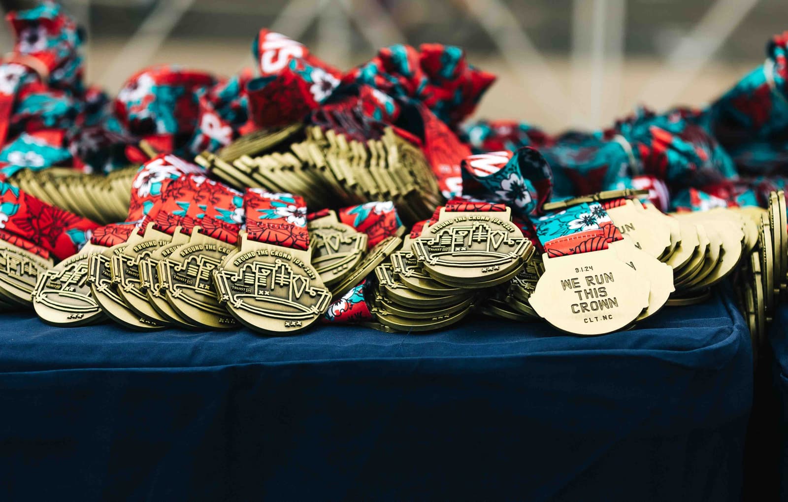 A table is covered with numerous gold medals featuring a city skyline design, each attached to a colorful red and blue ribbon. The medals read "We Run This Crown, CLT, NC.
