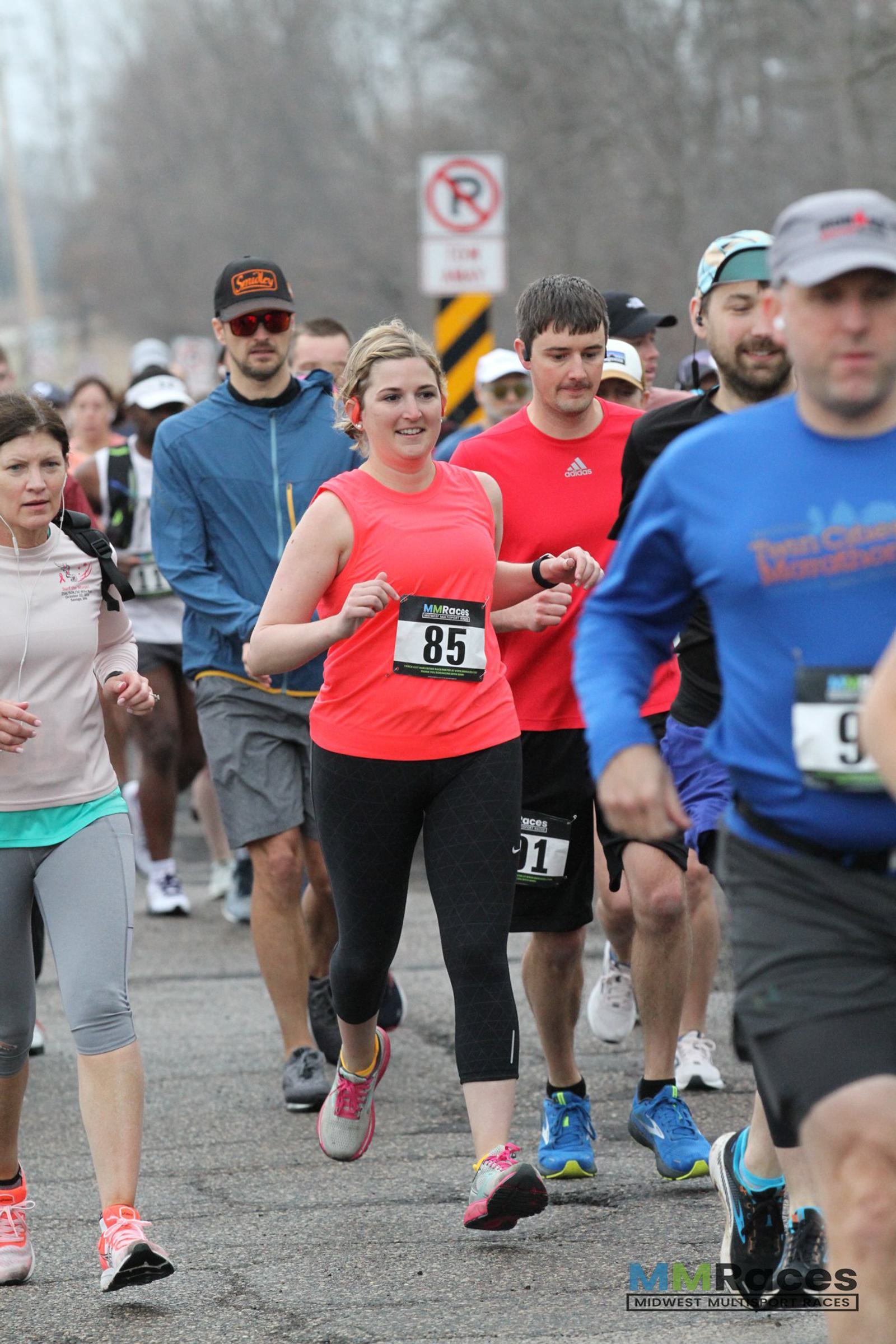 A group of runners participating in a road race. A woman wearing a pink top and bib number 85 is smiling in the center, surrounded by other runners in athletic gear on a cloudy day.