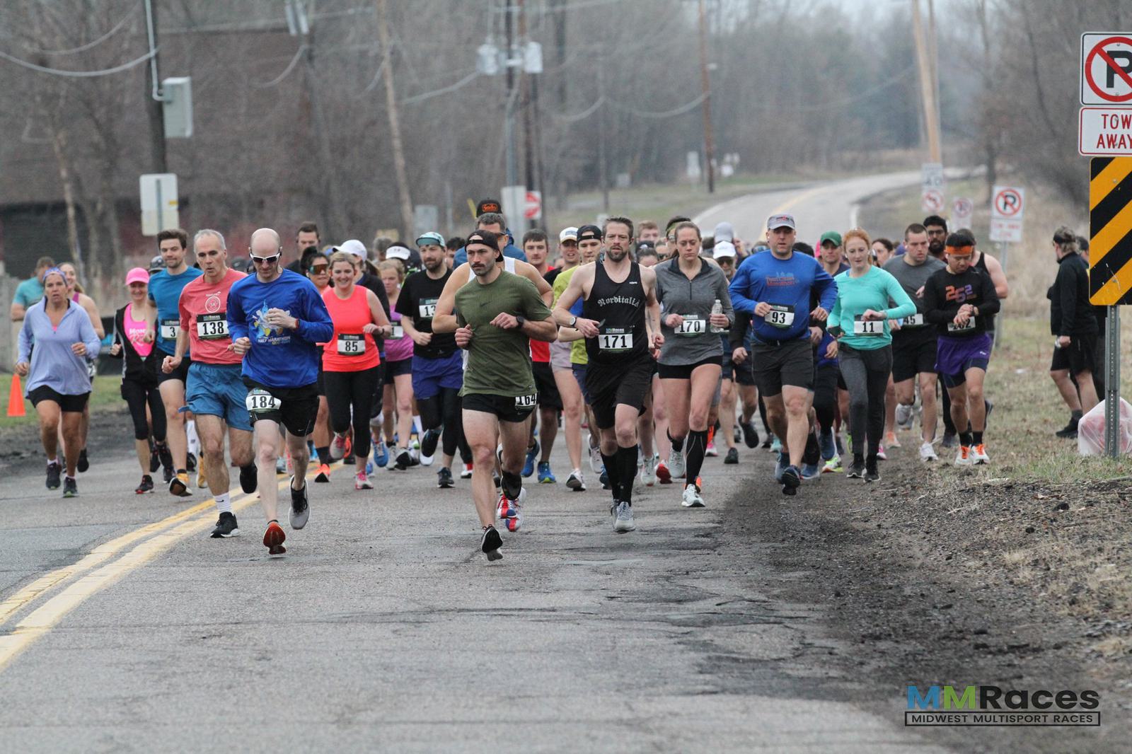 A large group of runners participating in a race on a rural road, wearing numbered bibs and athletic clothing. The weather appears overcast, and trees without leaves border the road. The MMR Races logo is visible.