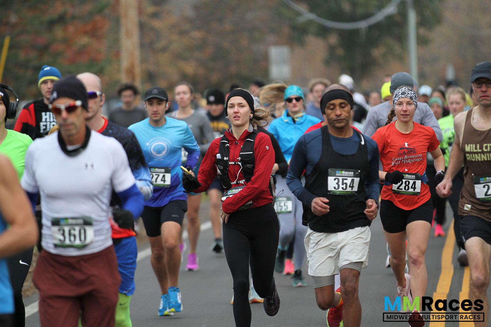 A group of runners, wearing race bibs and athletic clothing, participate in an outdoor road race on a cloudy day. Trees with autumn leaves line the street in the background. The MMRaces logo appears in the lower right corner.