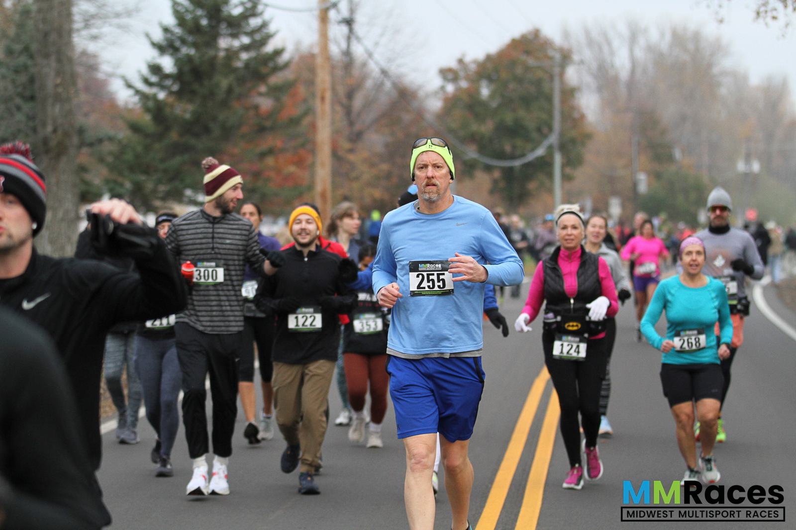A group of runners participates in a road race on an overcast fall day. The man in front wears a blue shirt, shorts, and a yellow headband. Other racers follow behind, and autumn trees line the road. The MM Races logo appears in the corner.