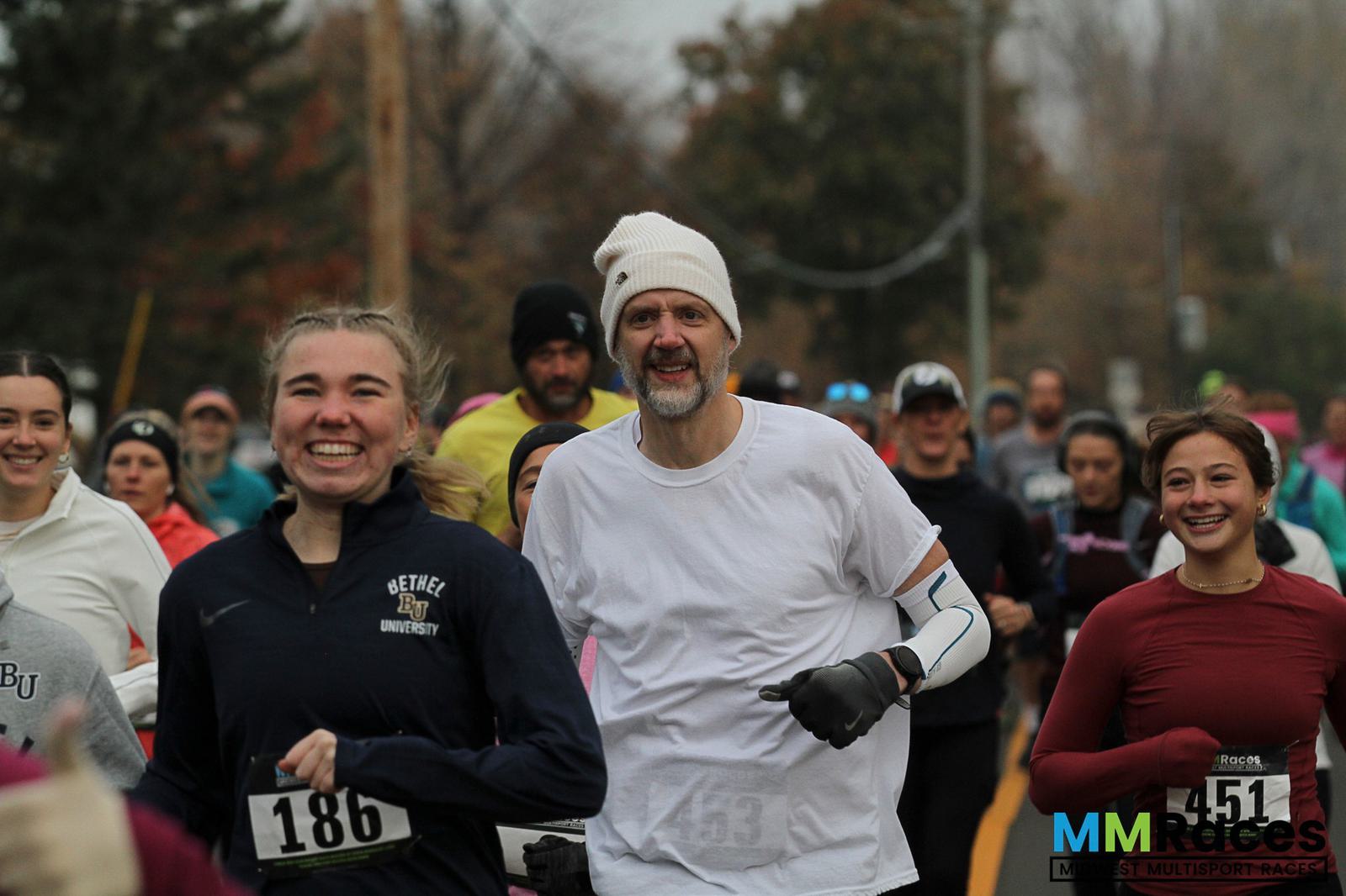 A group of runners, smiling and jogging outdoors during a race. The foreground shows a young woman in navy and a man in a white hat and shirt. Other participants run behind them on a cloudy day.