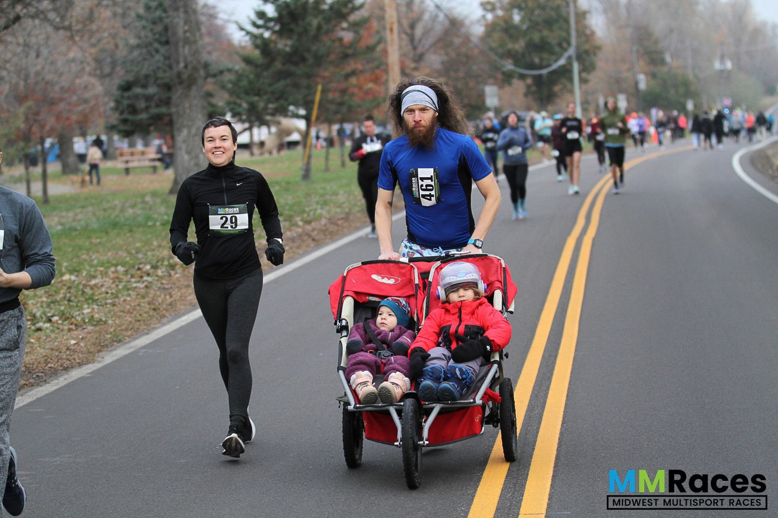 A man pushes a double stroller with two young children as he runs alongside a smiling woman during a road race. Other runners are visible in the background. The road is lined with trees. "MM Races" is visible in the corner.