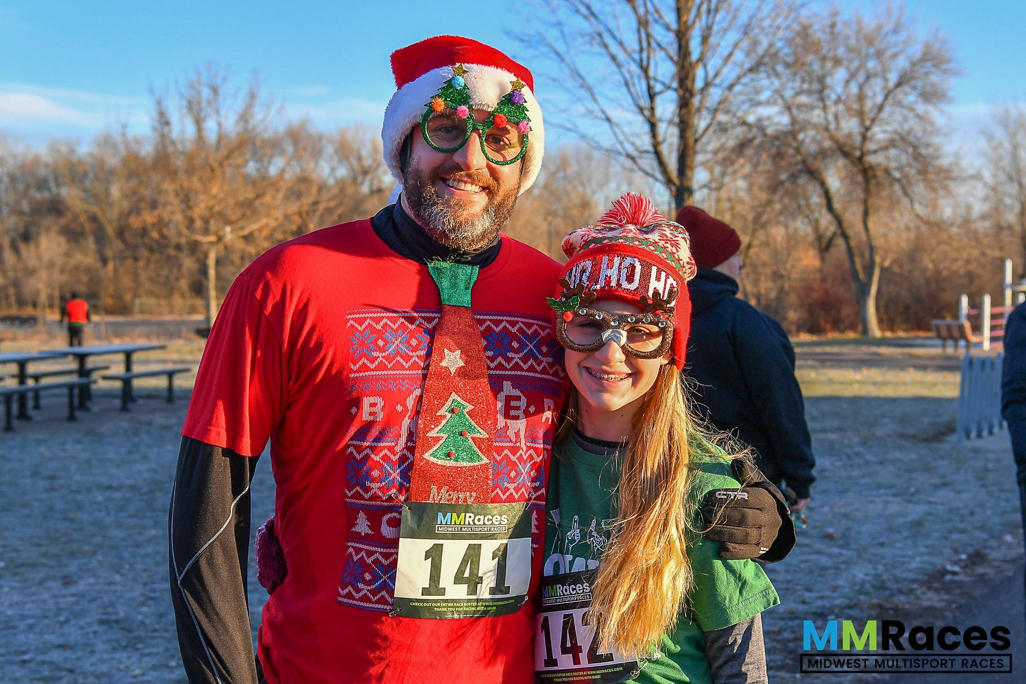 Two runners dressed in festive holiday attire, including Christmas hats and themed glasses, smile at the camera before a race outdoors on a sunny, winter day. The MM Races logo is visible in the corner.