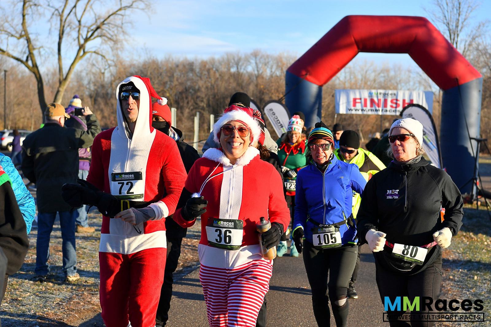 A group of runners in festive outfits, including holiday pajamas and costumes, approaches the finish line at an outdoor race event on a sunny day. The inflatable finish arch and race bibs are visible.