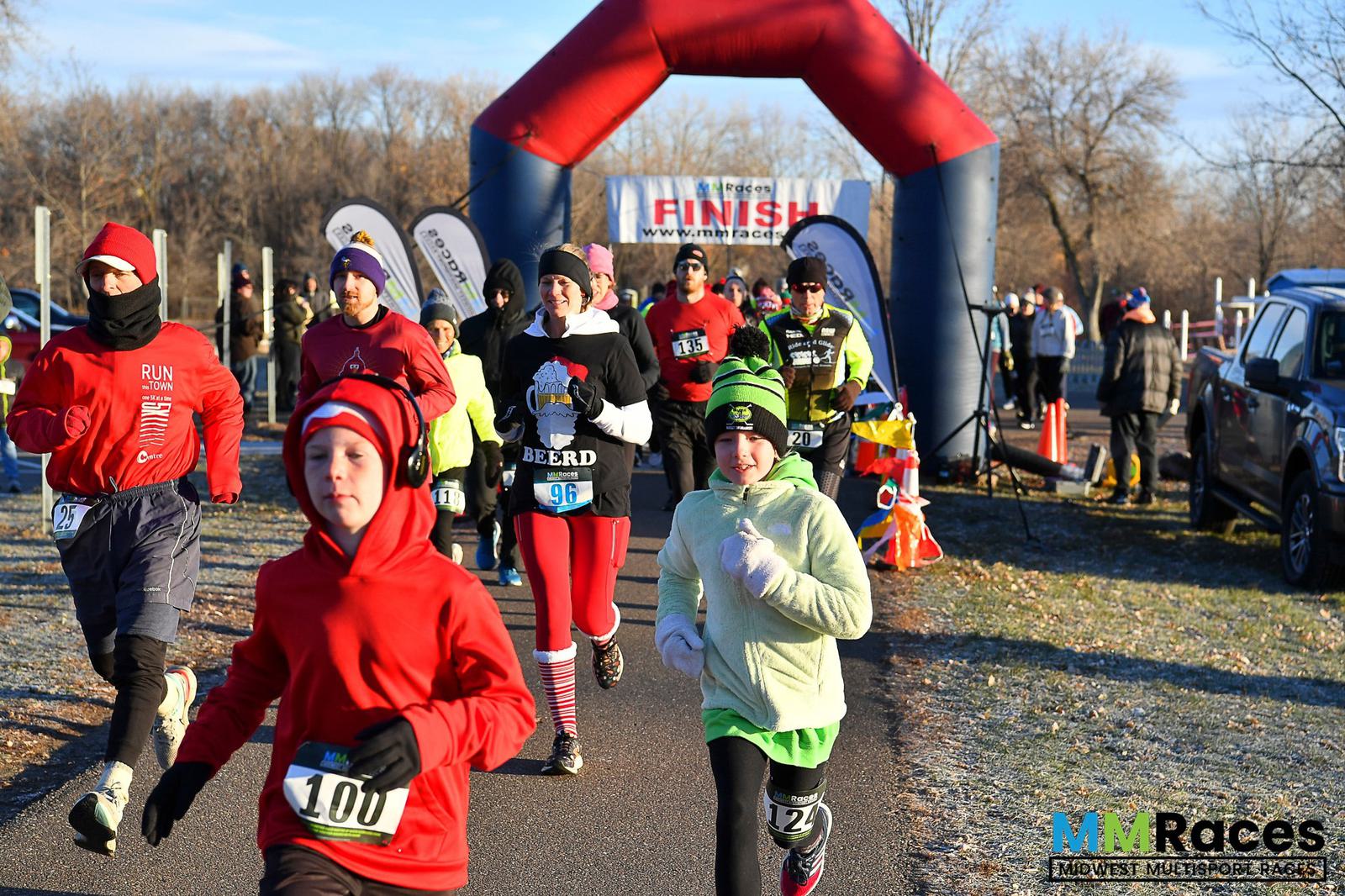 A group of runners, dressed in winter gear, participate in a race on a sunny day. An inflatable arch with "FINISH" written on it stands in the background. The Midwest Multisport Races logo is in the bottom right corner.