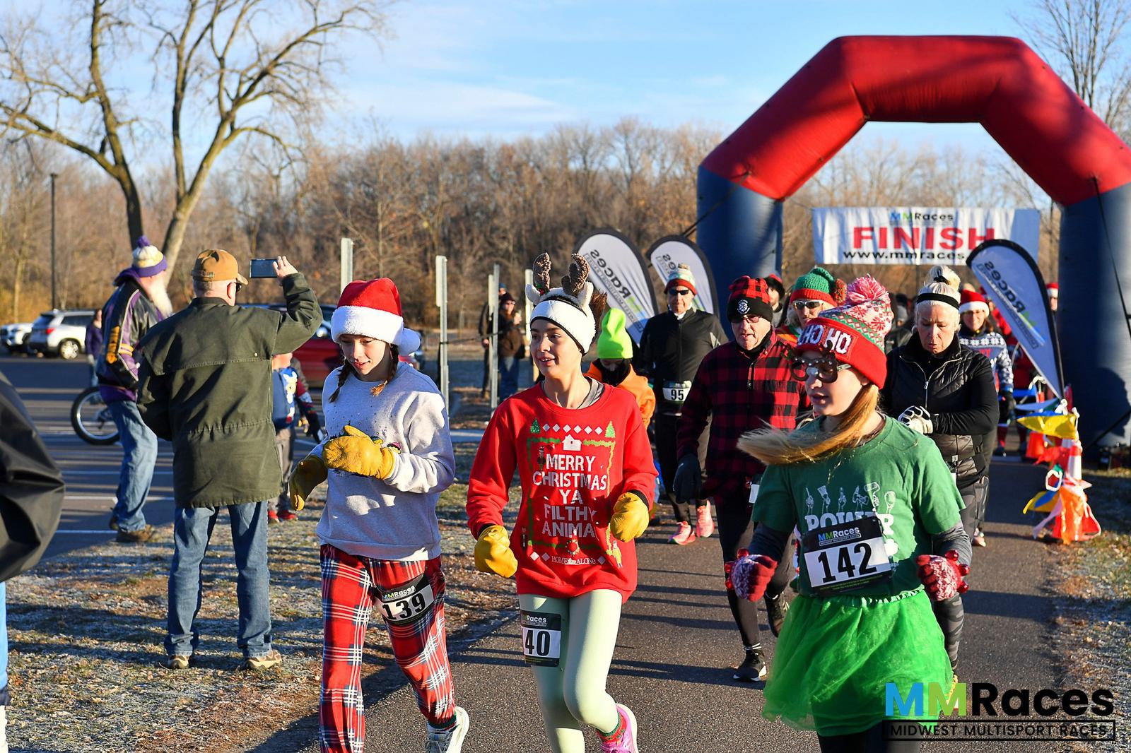 Runners in festive holiday outfits, including Santa hats and Christmas sweaters, approach the finish line at an outdoor winter race, with a red and blue inflatable arch and spectators in the background.