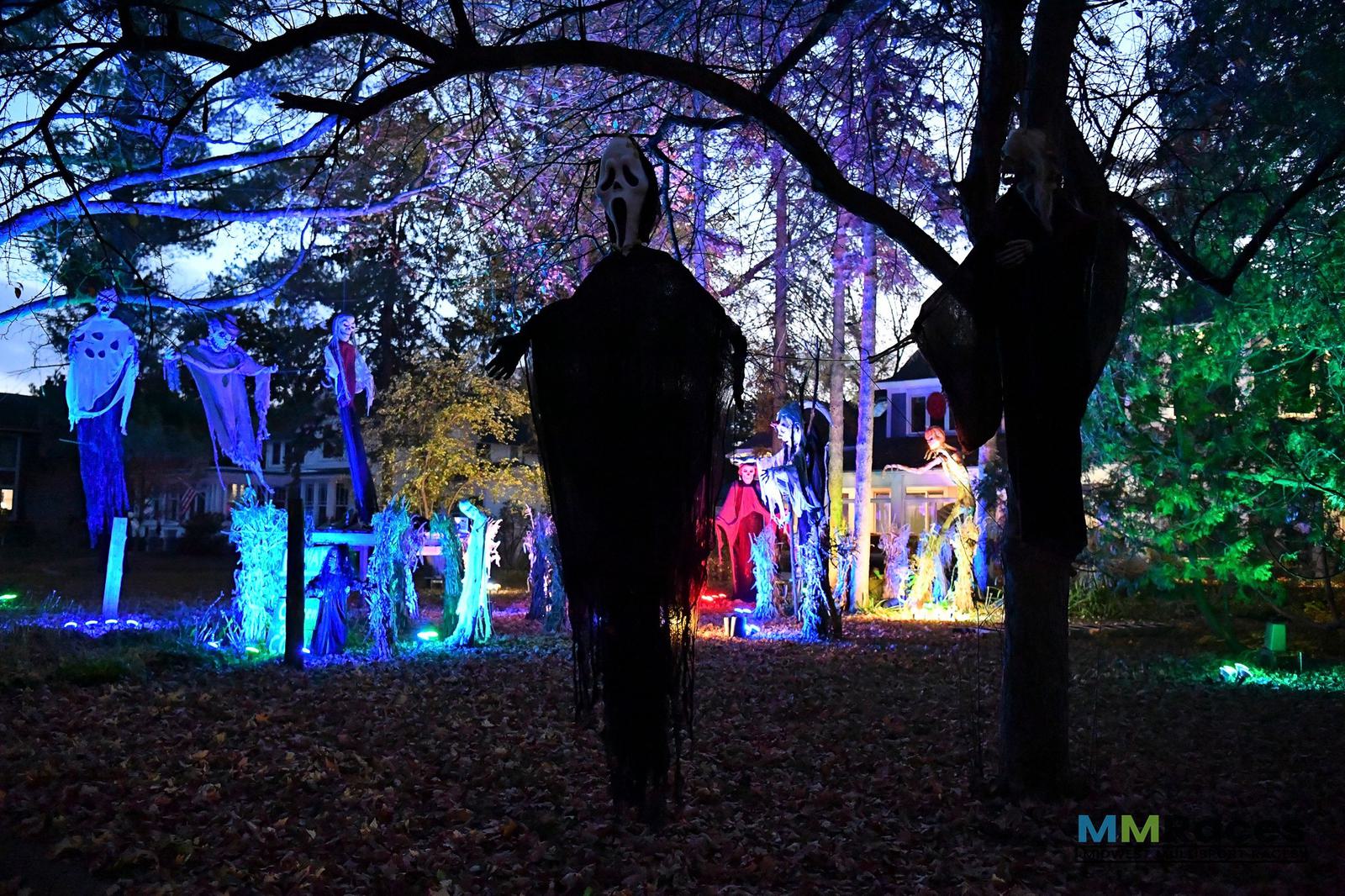 Halloween decorations in a yard at dusk feature glowing lights, hanging ghost and grim reaper figures, and spooky props among trees and fallen leaves, creating a colorful and eerie scene.