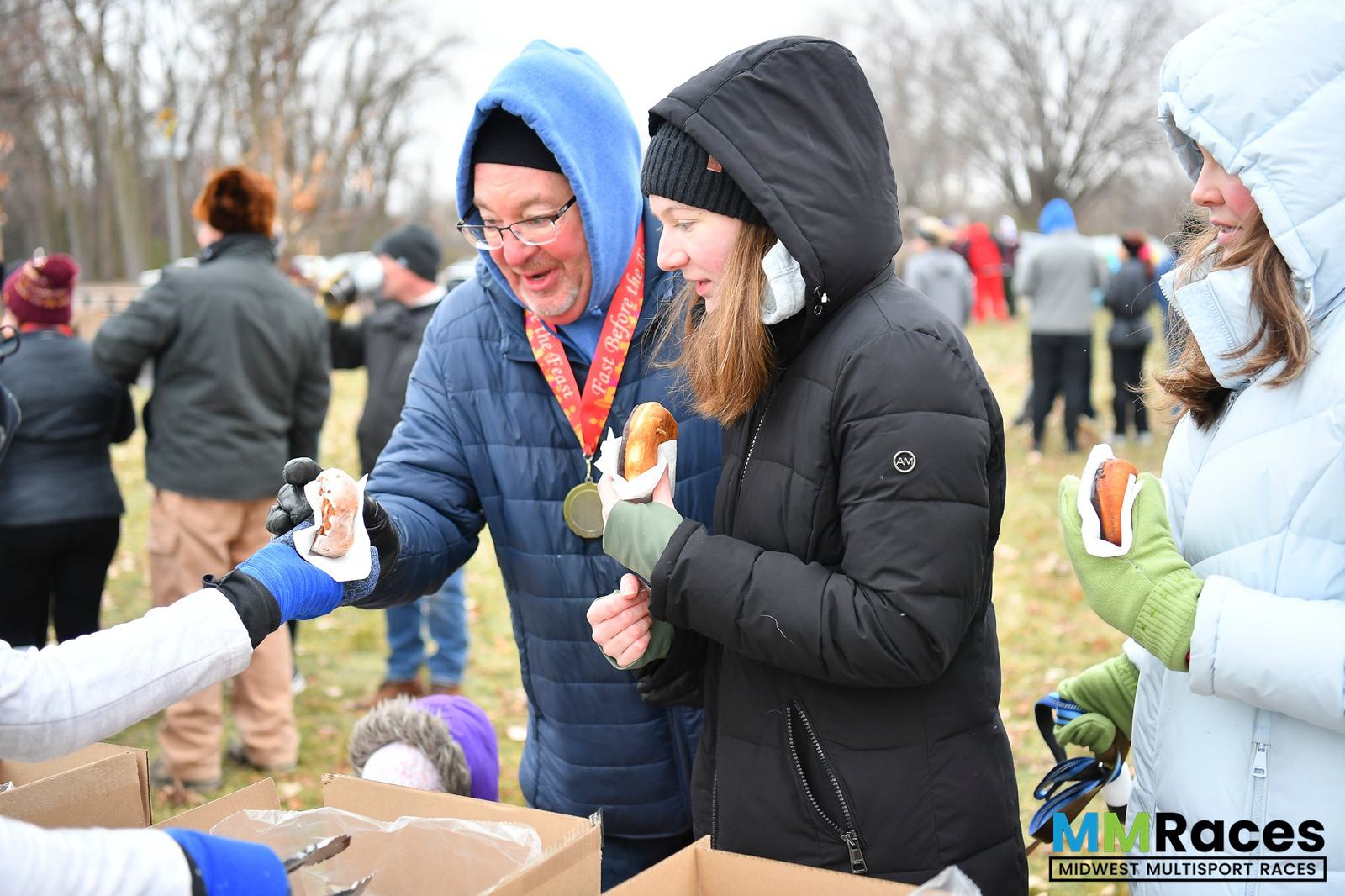 Two people in winter jackets and hats smile as they receive hot dogs from gloved volunteers at an outdoor event. Other participants and trees are visible in the background. The MM Races logo is in the corner.