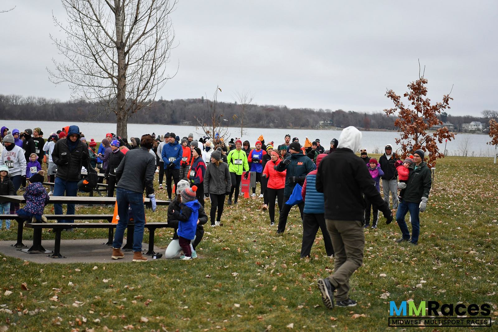 A group of people dressed in warm clothing gather at a park near a lake on a cloudy day, preparing for a race event. Some stand near picnic tables, while others walk on the grass. The Midwest Multisport Races logo is visible.