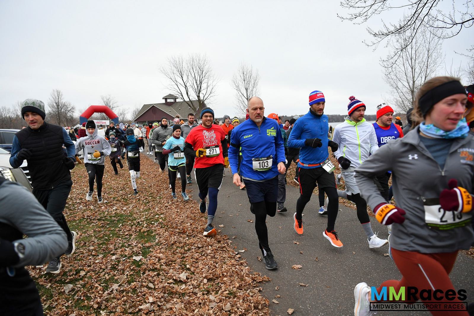 A group of runners dressed in winter athletic gear participates in an outdoor race on a paved path bordered by fallen leaves, with bare trees and an overcast sky in the background.