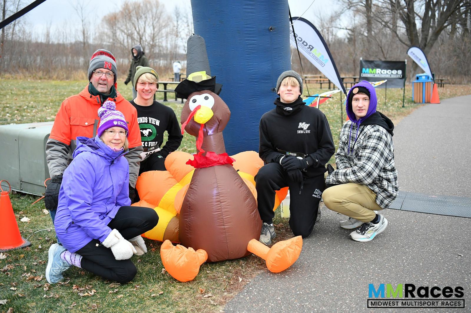 Five people in winter clothing pose around a large inflatable turkey at an outdoor event. The ground is paved, and there are banners in the background for "MMRaces: Midwest Multisport Races.