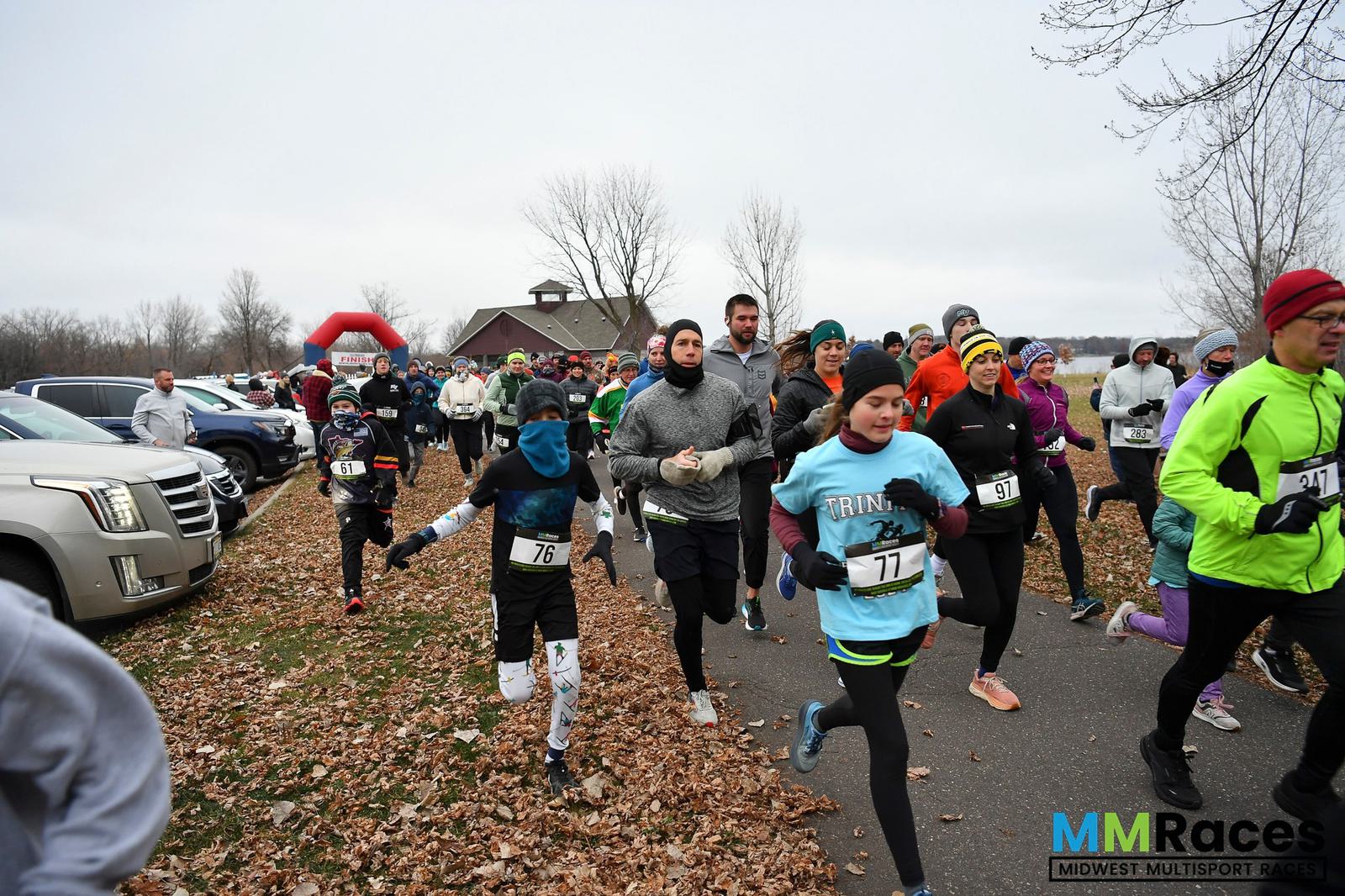 A group of runners bundled in winter clothing participate in a race on a leaf-covered path near parked cars, with bare trees and cloudy skies in the background. The MM Races logo is visible in the bottom right corner.