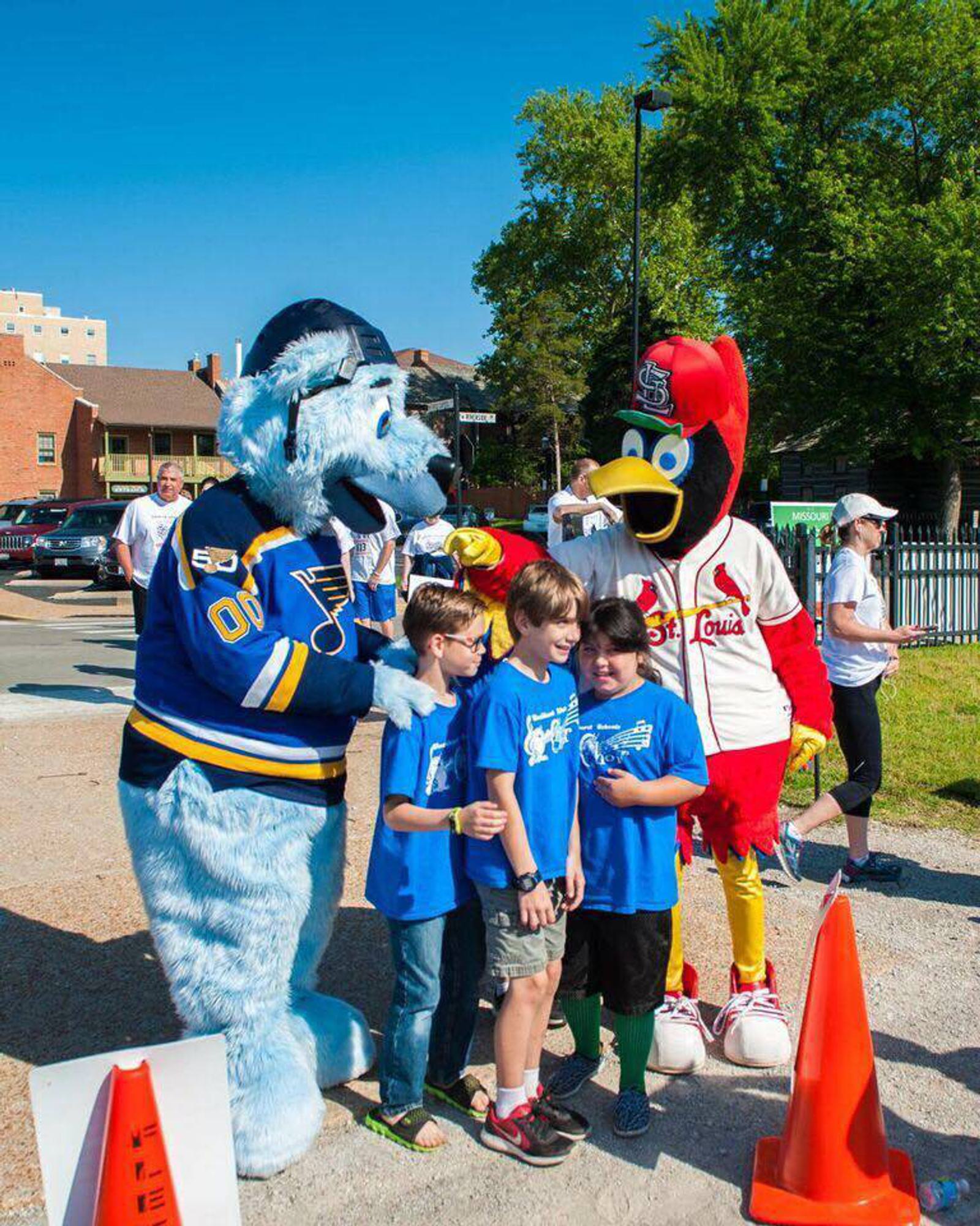 A group of children in blue t-shirts pose outdoors with a white furry mascot in a hockey jersey and a red bird mascot in a baseball uniform. There are orange cones in the foreground and trees and buildings in the background.