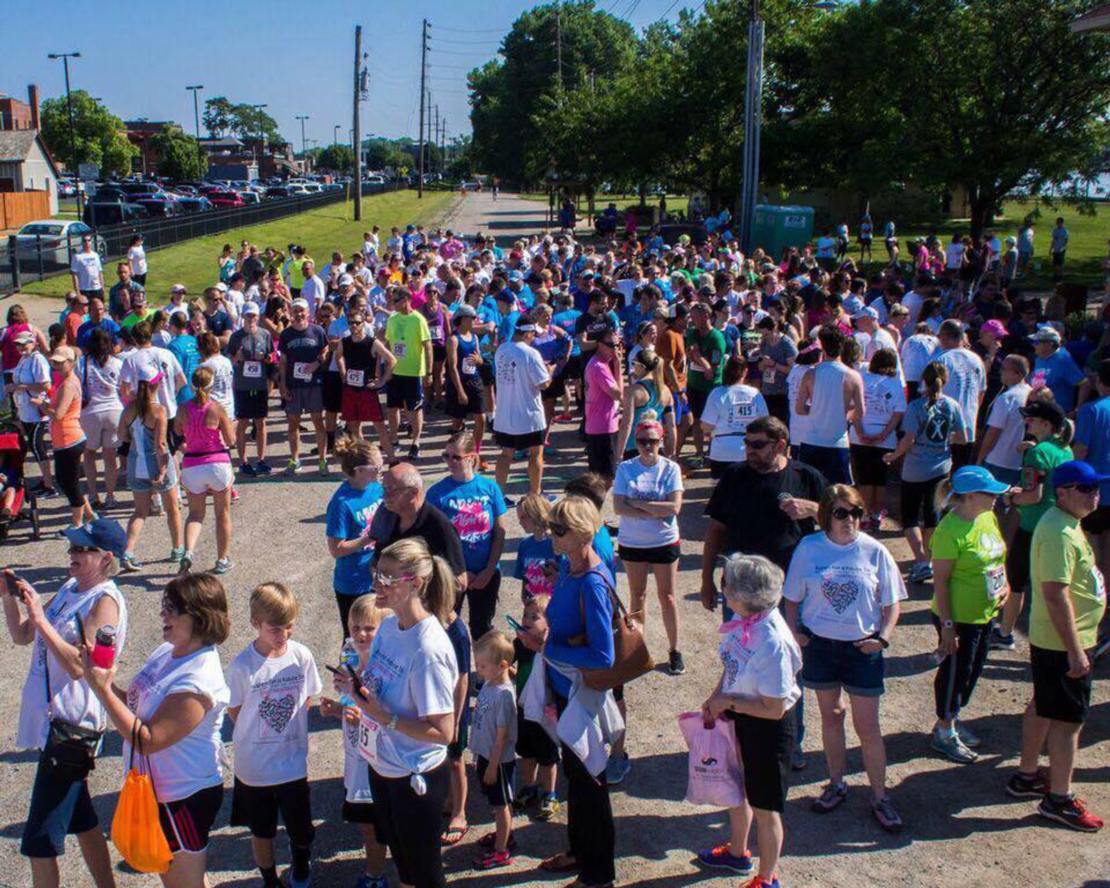 A large group of people of various ages gather at the starting area of an outdoor fun run or charity race, wearing numbered bibs and casual athletic clothing. The sunny day shows some greenery and parked cars in the background.