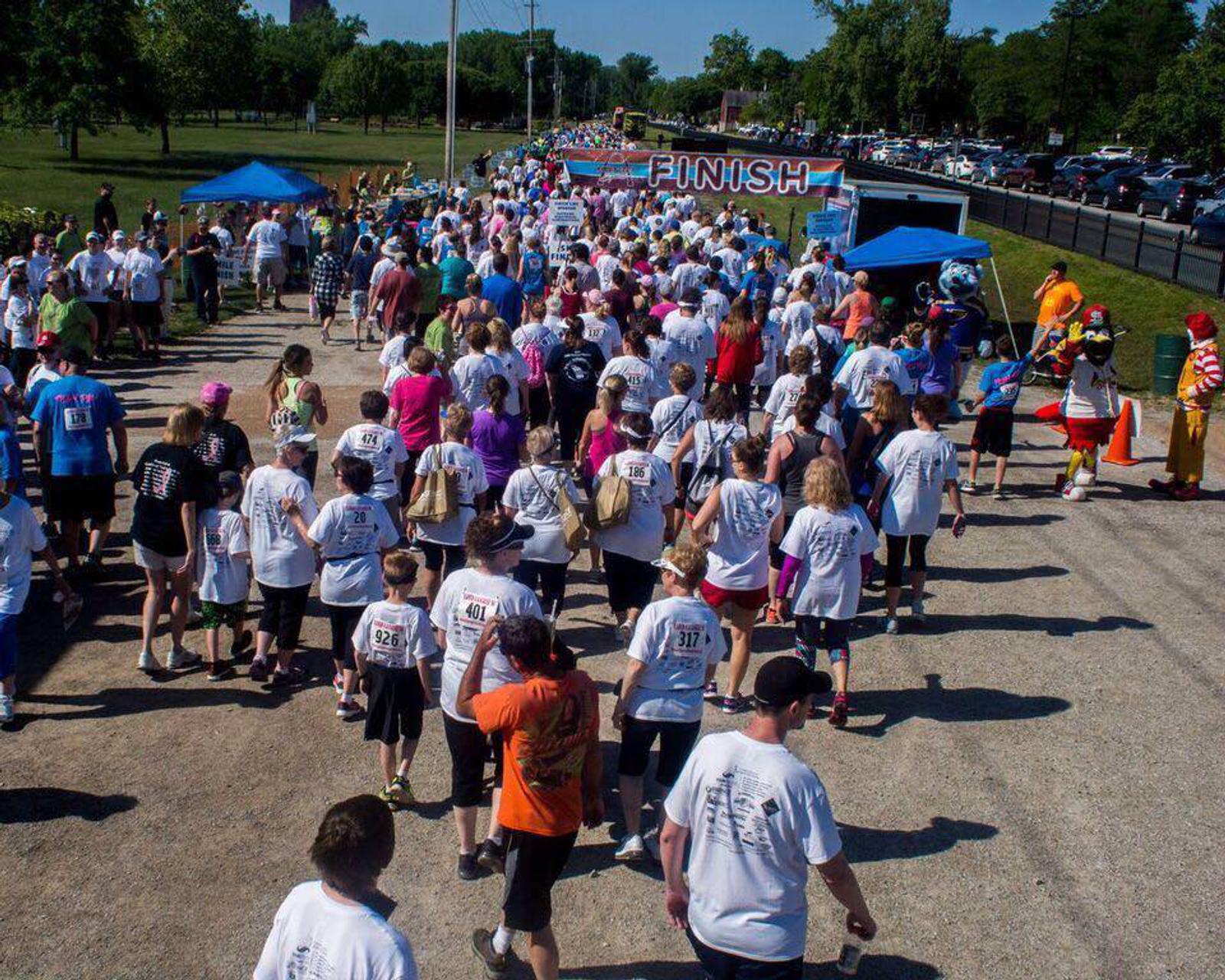 A large group of people in athletic gear participate in a fun run or charity walk. They are approaching a finish line banner. The event takes place outdoors on a sunny day, with trees and parked cars visible in the background.