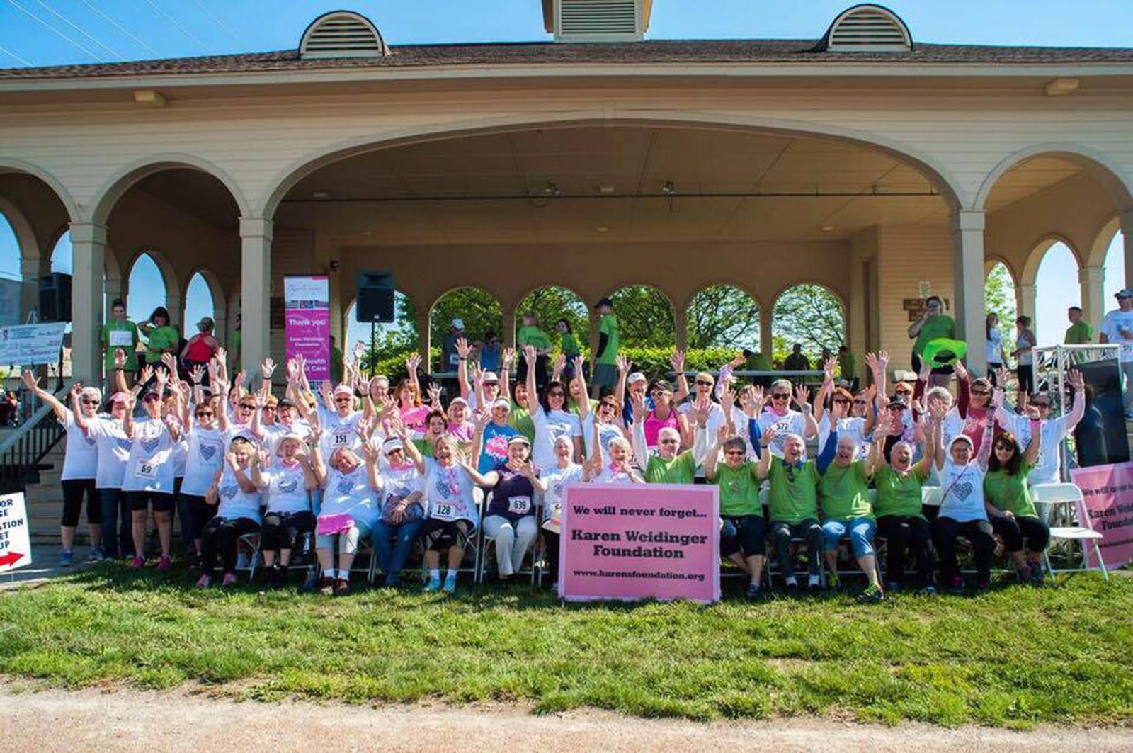 A group of people gather in front of a pavilion wearing pink and green shirts, holding signs for the Karen Weidinger Foundation. They are raising their arms in celebration on a sunny day.