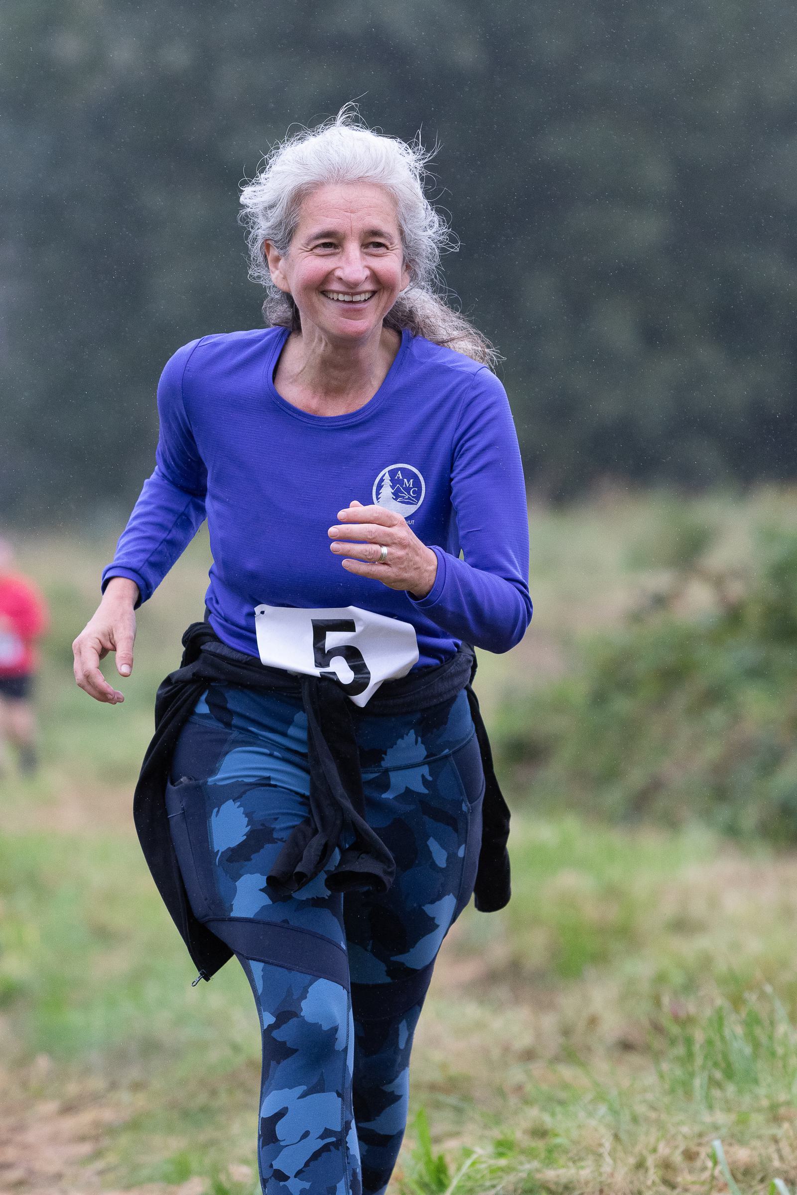 An older woman with long gray hair is running outdoors. She is wearing a blue long-sleeve shirt and camouflage-patterned leggings, with a race number 5 on her waist. The background is grassy and blurred. She appears to be smiling.