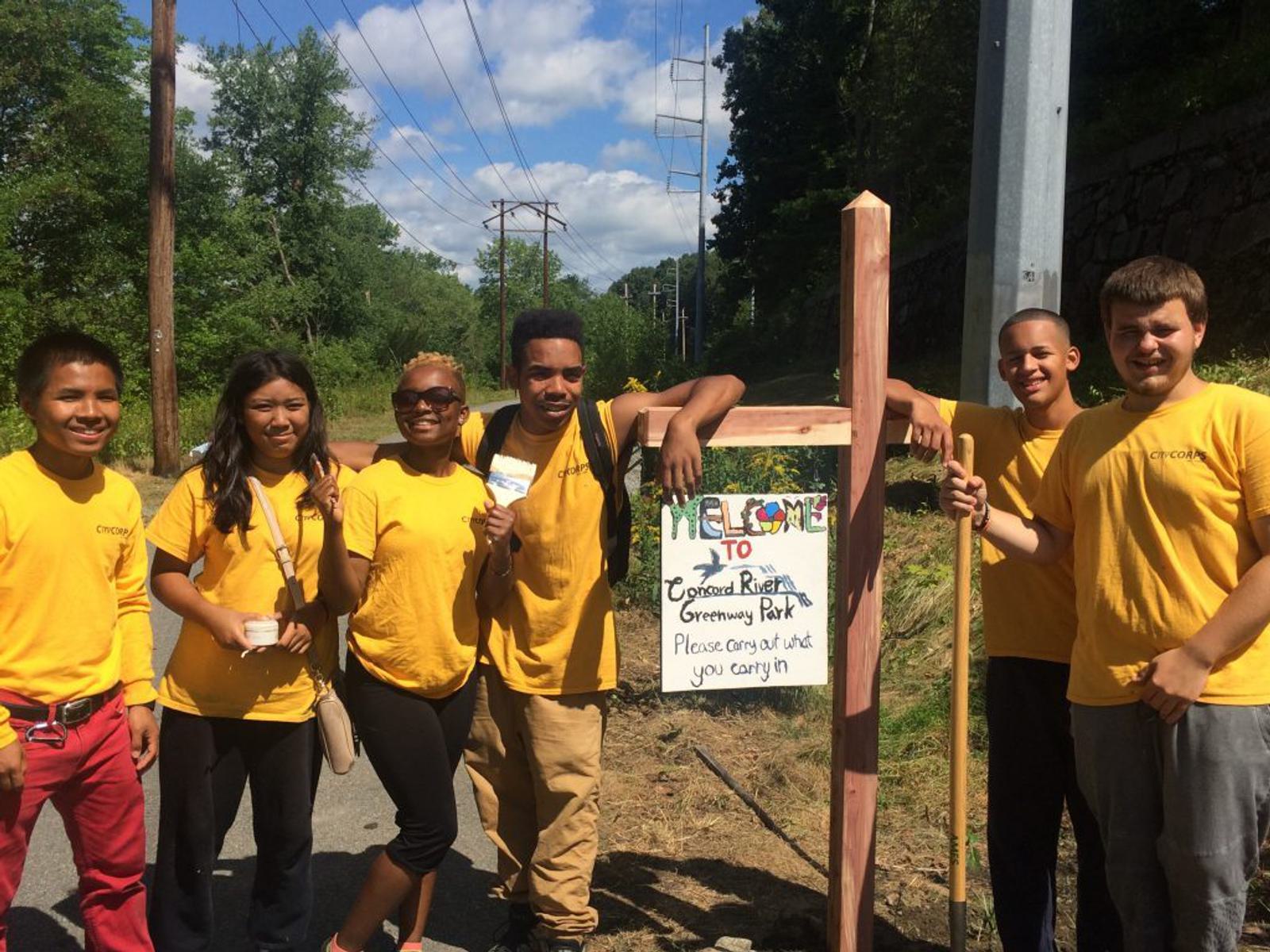 A group of six young adults in yellow shirts stand smiling next to a sign saying "Welcome to Coyego River Greenway Park. Please carry out what you carry in." They are outdoors with trees, power lines, and a trail in the background.