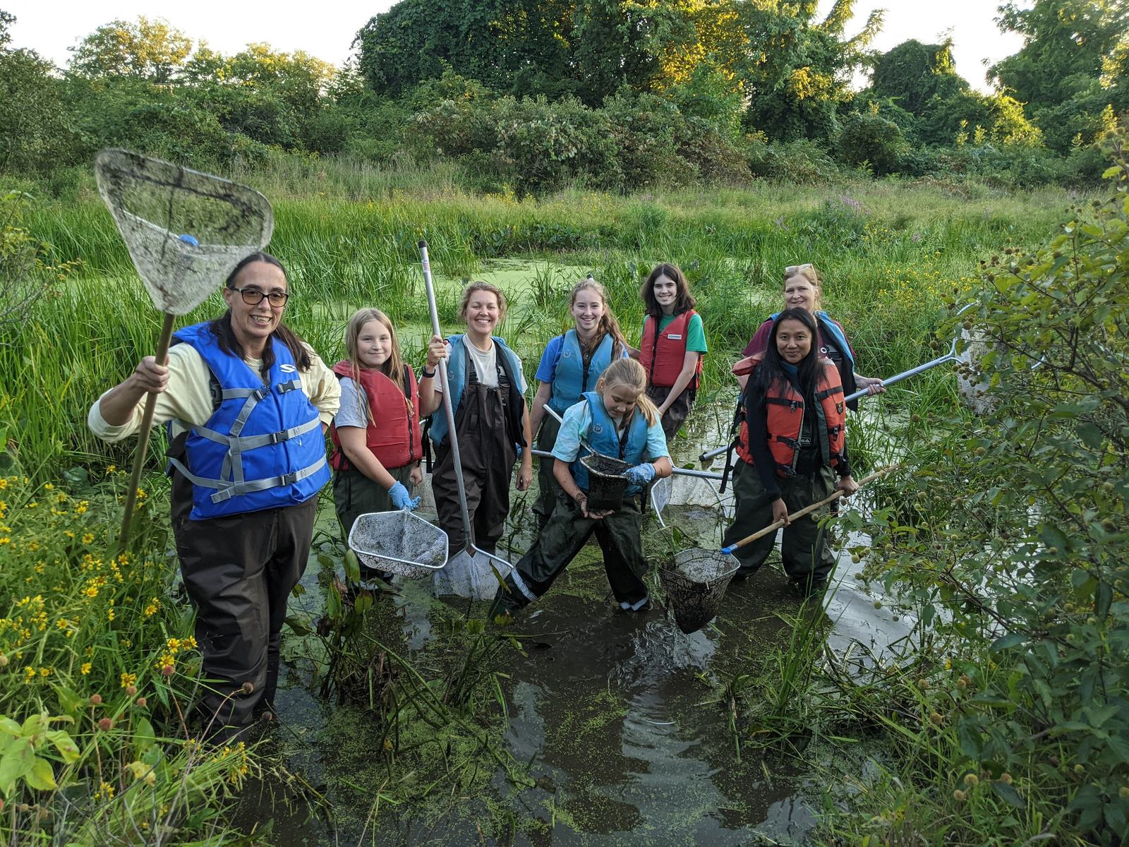 A group of eight people standing in a shallow pond surrounded by greenery, holding nets and wearing waders and life jackets. They are smiling and appear to be engaged in an outdoor water activity or research.