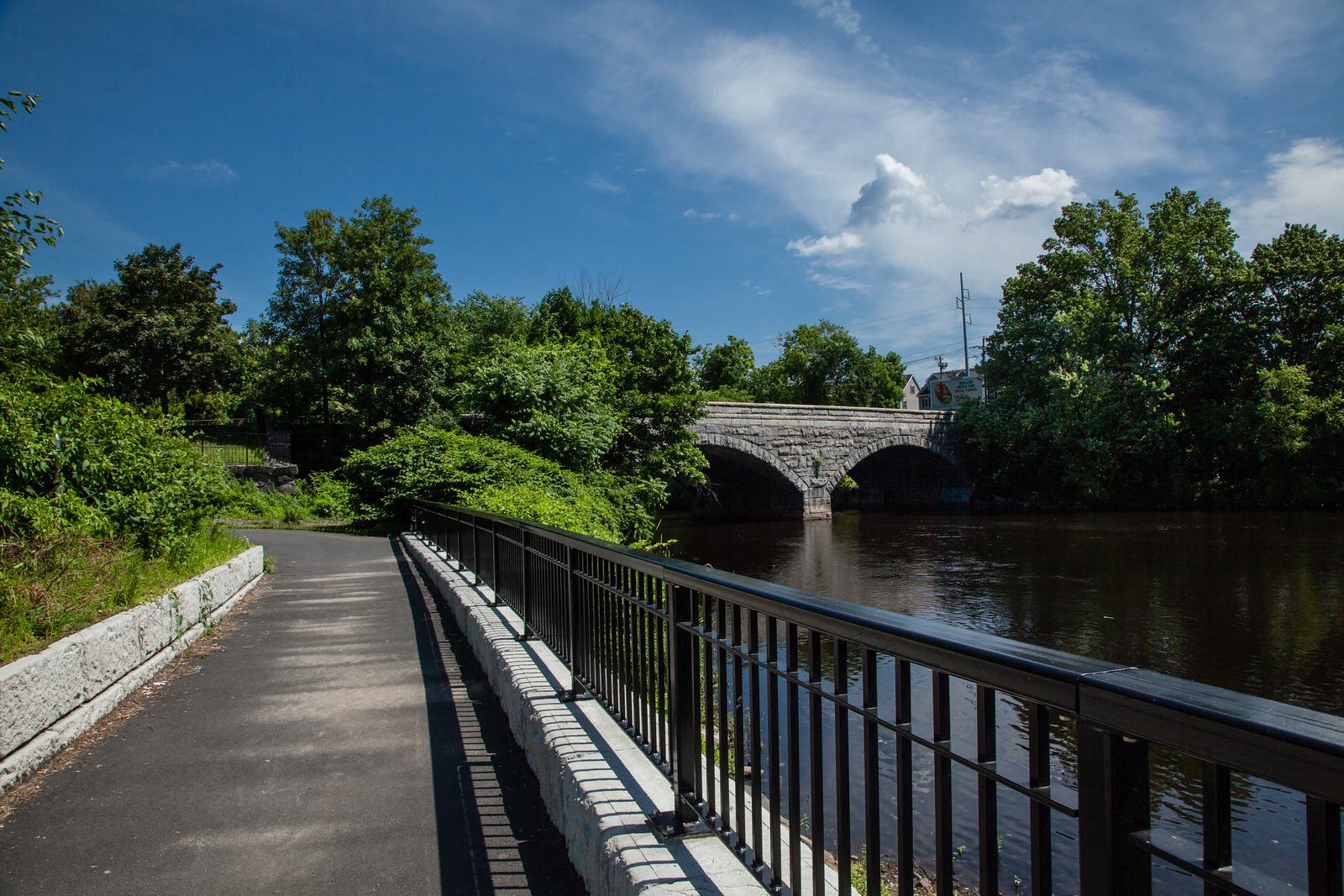 A paved pathway runs alongside a calm river, partially bordered by a railing on the right. A stone bridge spans the river in the background, surrounded by lush green trees under a partly cloudy blue sky.