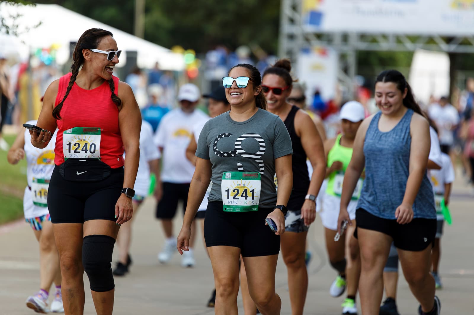 A group of smiling women wearing athletic clothes and bib numbers walk together at a race event, surrounded by other participants on a sunny day.