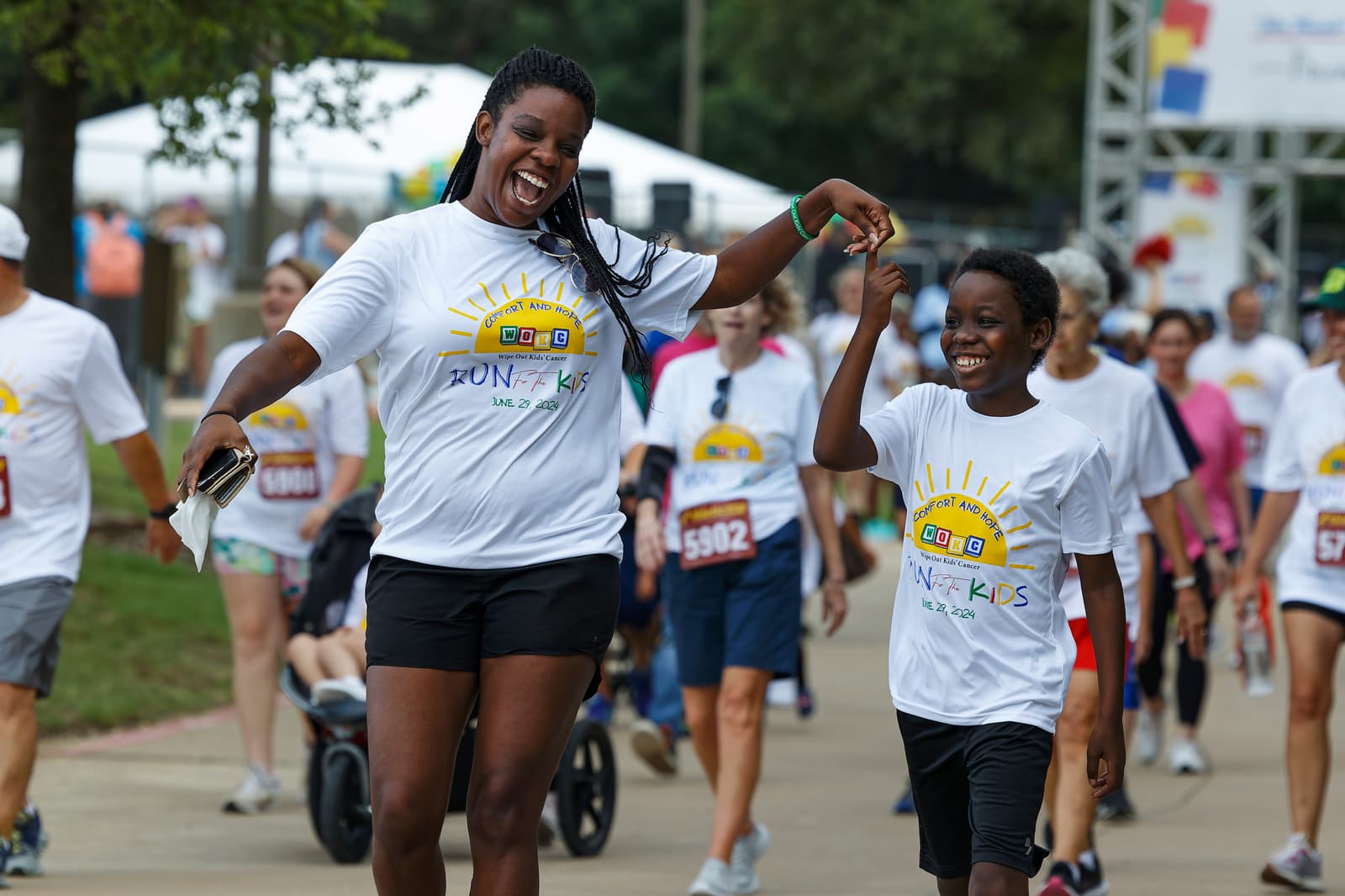A woman and a boy, both smiling and holding hands, participate in a charity run wearing matching "Run for Kids" T-shirts. Other runners and families are visible in the background.