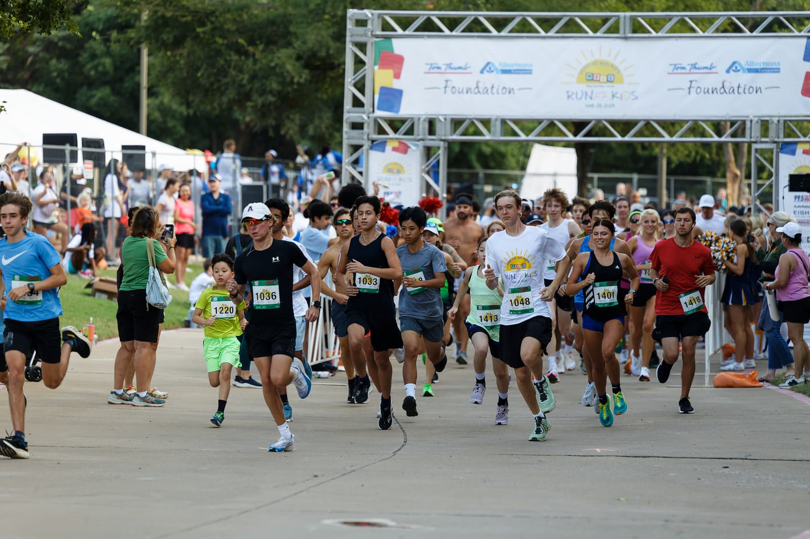 A diverse group of runners, including adults and children, start a race at an outdoor event. Spectators watch from the sides, and a banner reading "Foundation Run" hangs above the starting line.