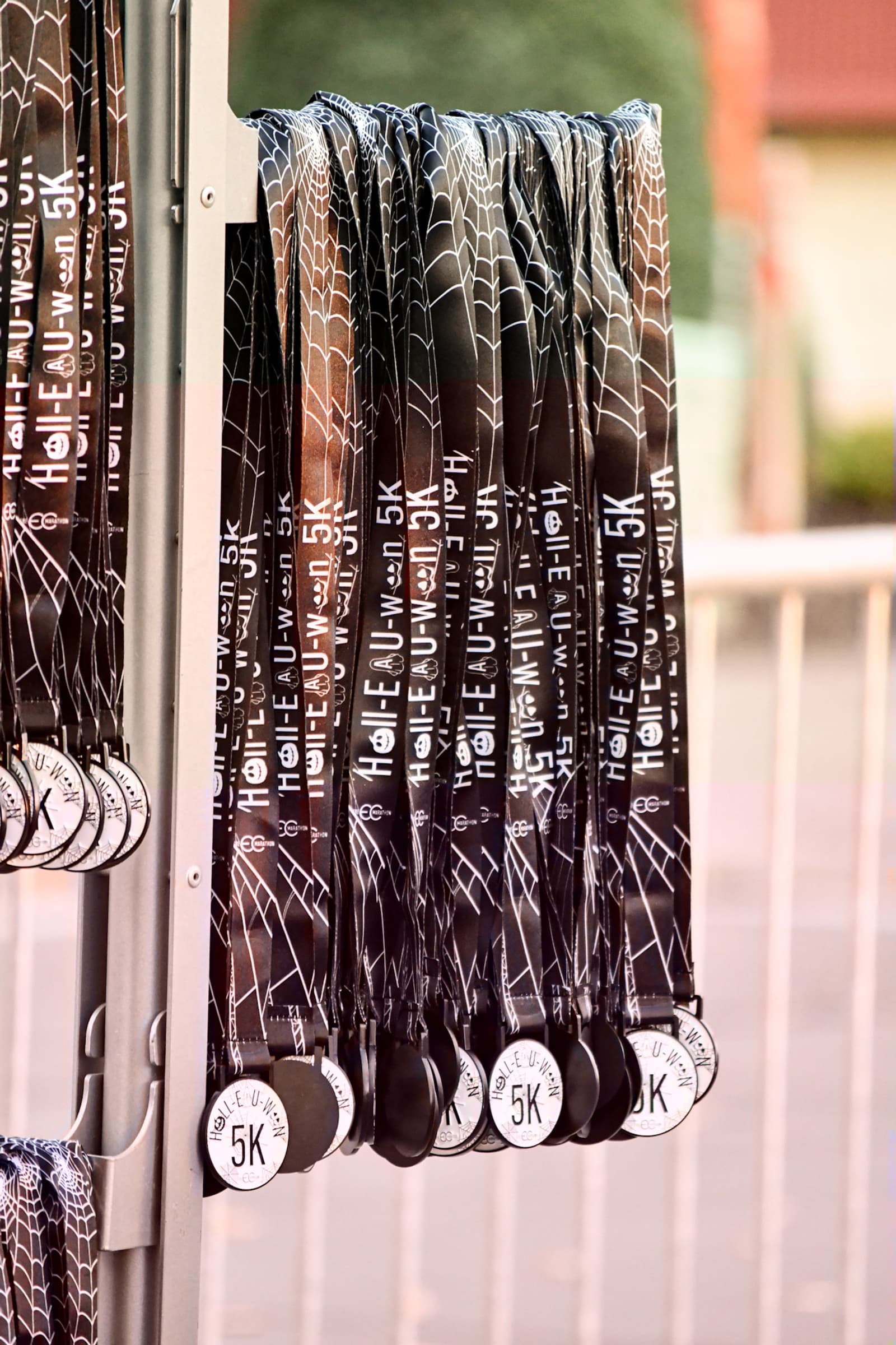 Several black and white finisher medals for a 5K event hang from a rack, each with a ribbon displaying the text "Haunt Our Town 5K." The background is slightly blurred.