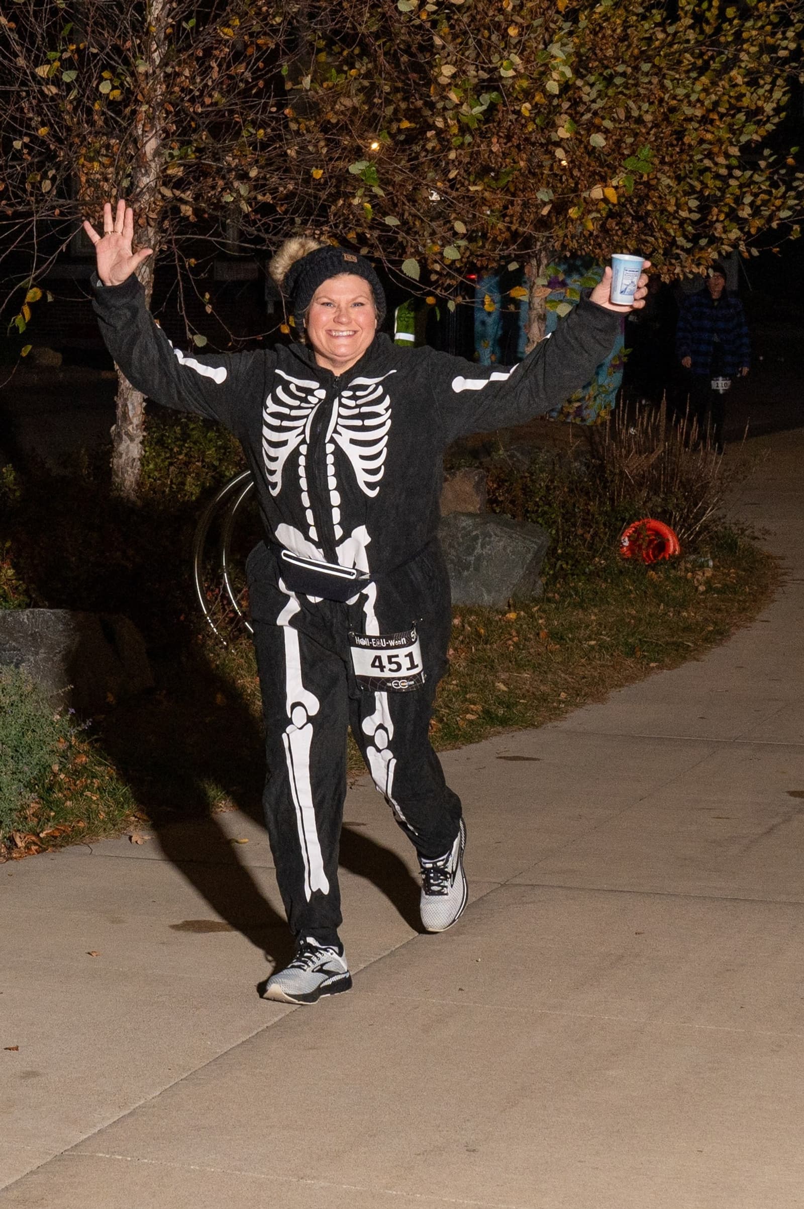 A person wearing a skeleton costume, race bib 451, and running shoes runs on a sidewalk at night, smiling and holding a cup, with autumn trees and leaves in the background.