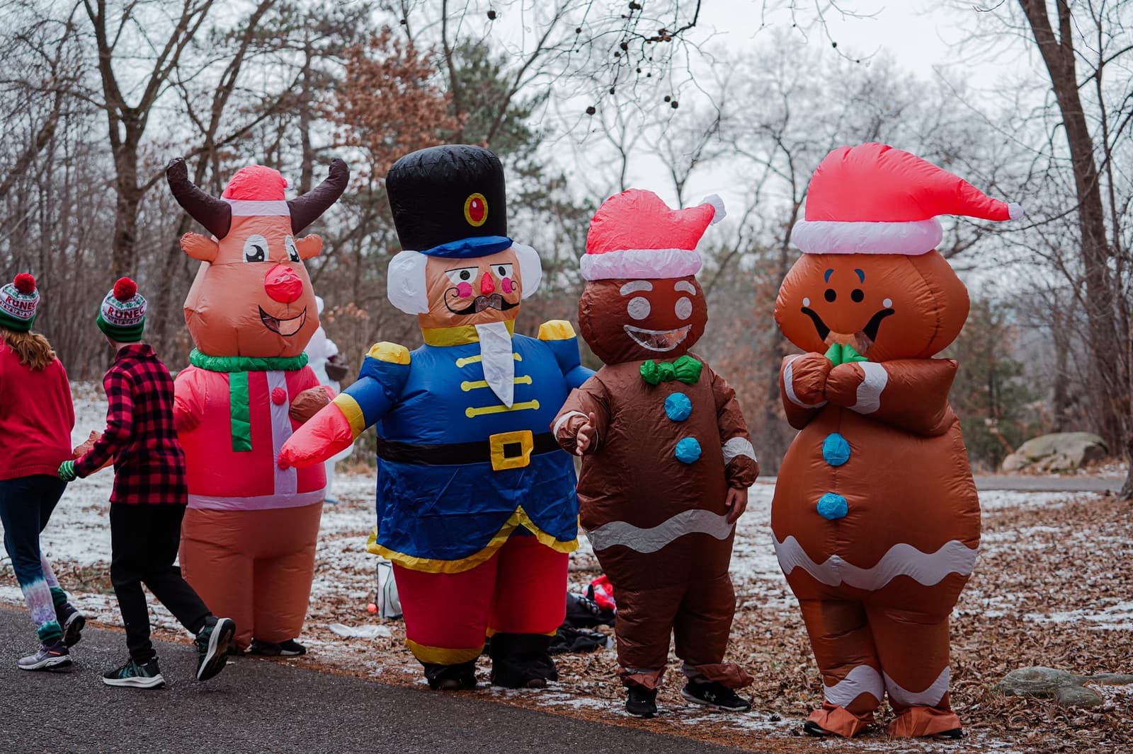 Four people in inflatable holiday costumes—reindeer, nutcracker, gingerbread man, and gingerbread woman—stand outdoors on a winter day, while two others in festive hats walk past on a paved path.