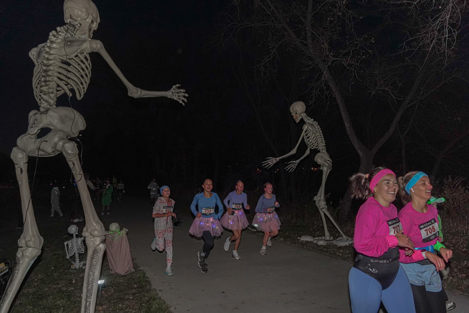 A group of runners, some in tutus and race bibs, jog down a path at night, smiling as they pass two large, posed skeleton decorations and bare trees.