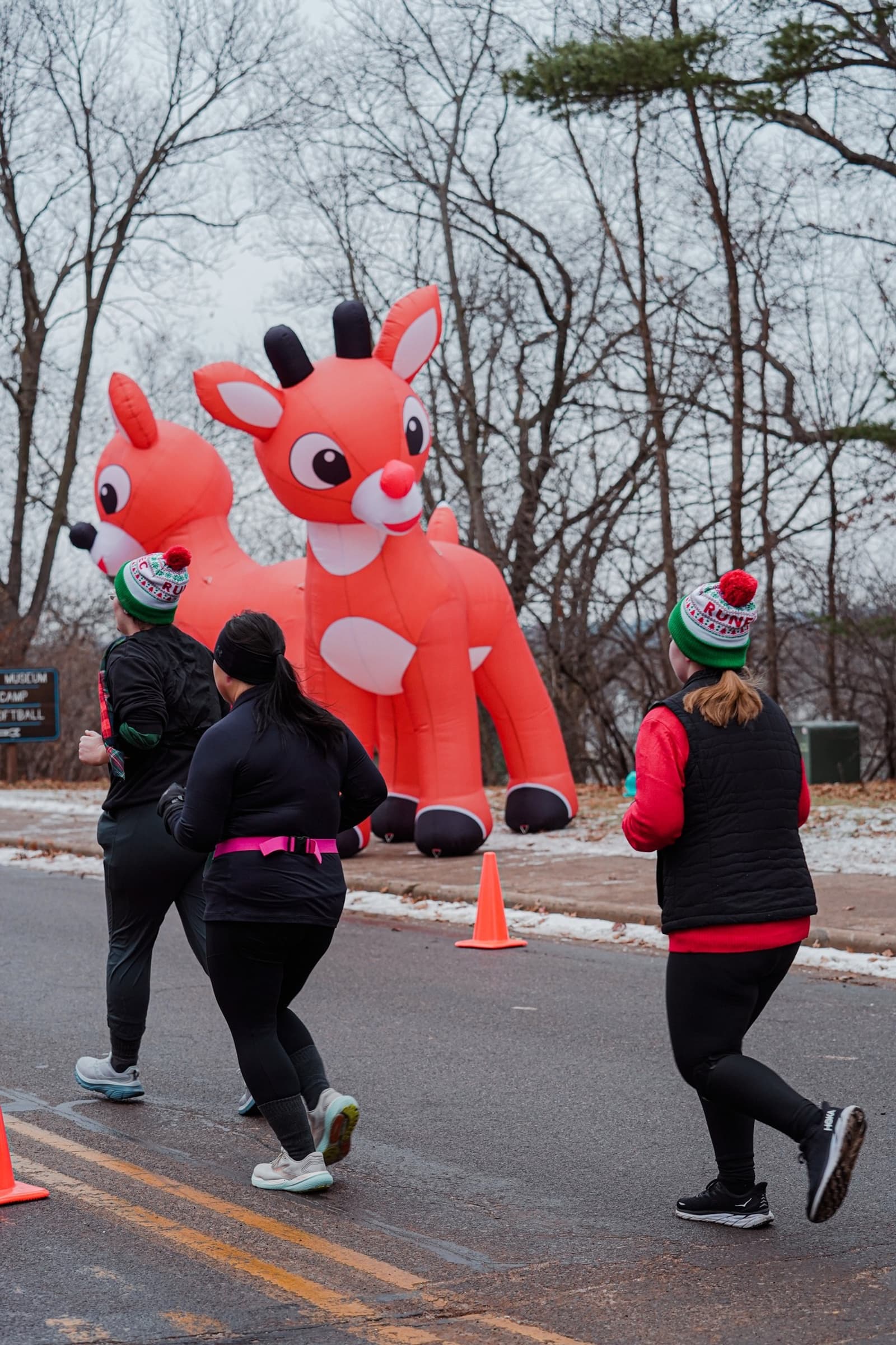 Three people in winter clothing run on a road past large inflatable reindeer decorations. Leafless trees and orange cones are visible in the background, creating a festive outdoor scene.