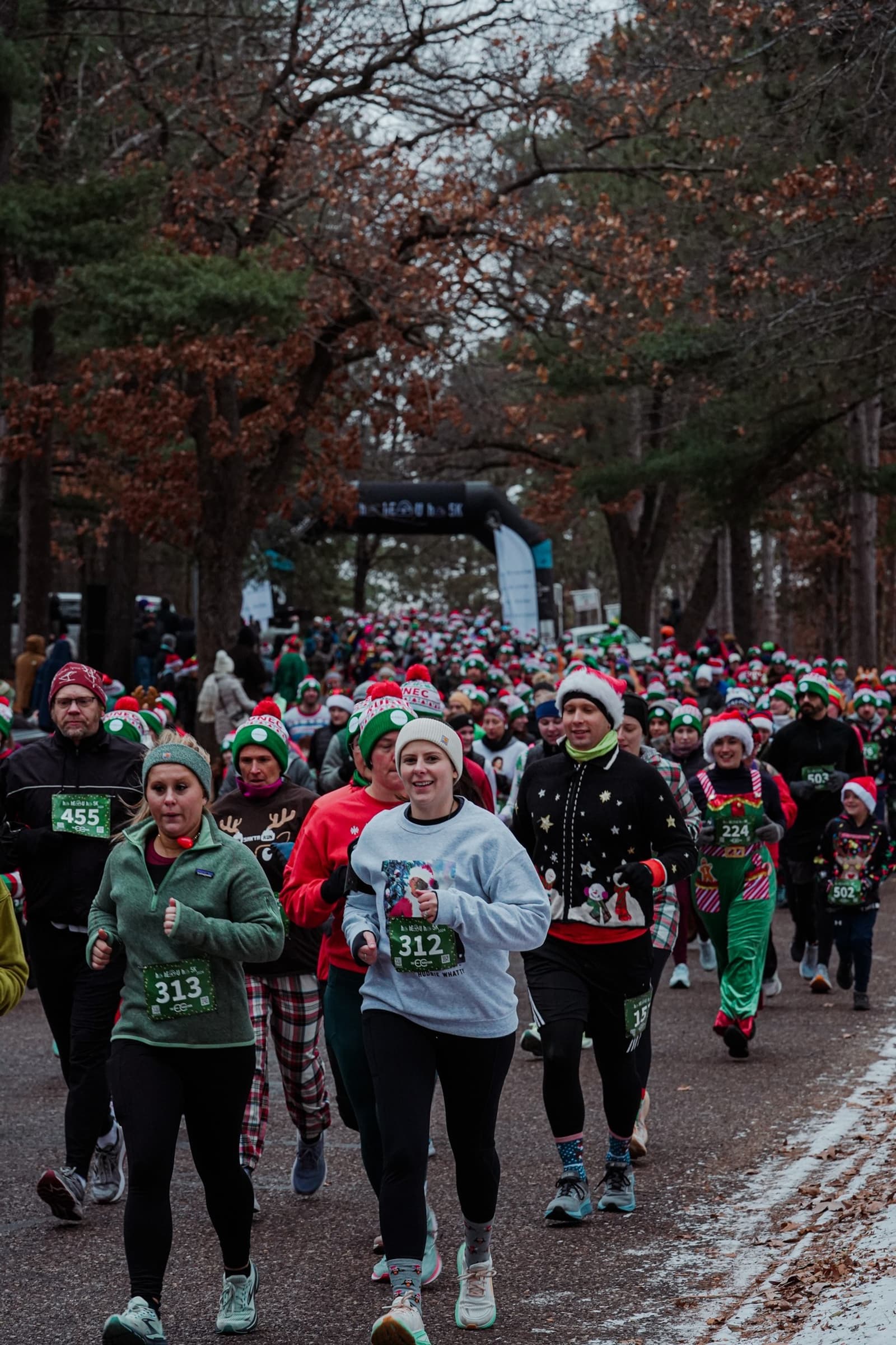 A large group of runners, many wearing festive holiday attire and Santa hats, participate in a race on a tree-lined road during winter. The atmosphere is lively and cheerful.