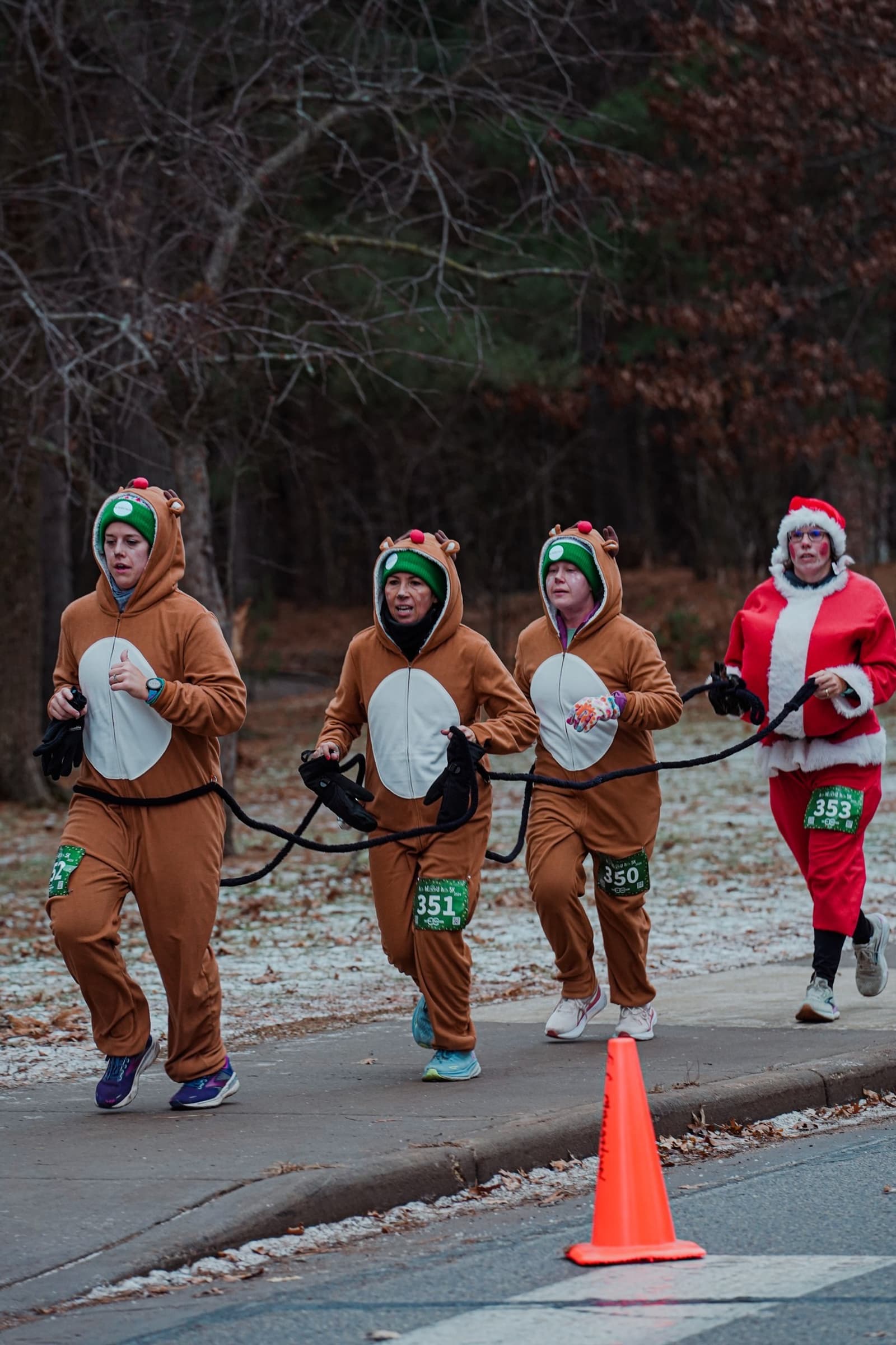 Four people are running outdoors in costume; three are dressed as reindeer and one as Santa Claus. They are connected by a rope and running on a path beside a road with an orange traffic cone visible.