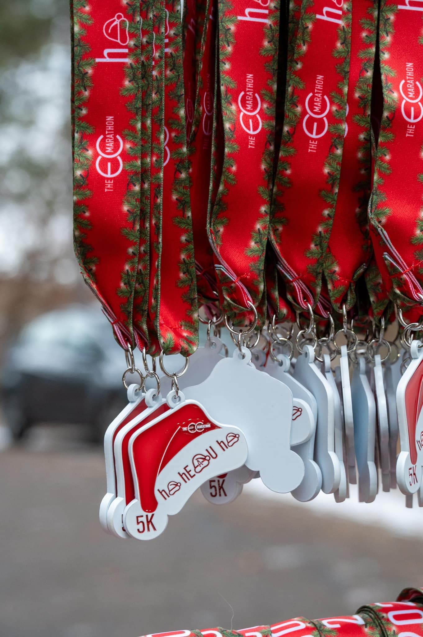 A close-up of several holiday-themed medals with red and green ribbons hanging on hooks. The medals are shaped like Santa hats and labeled “5K.” A blurred outdoor background is visible.
