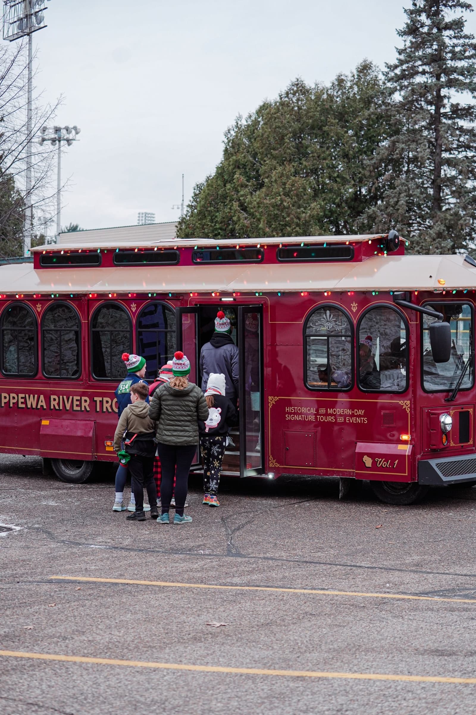 A group of people wearing winter clothing and festive hats stand near the open door of a red trolley with "Chippewa River Trolley" written on the side. Trees and a parking lot are visible in the background.