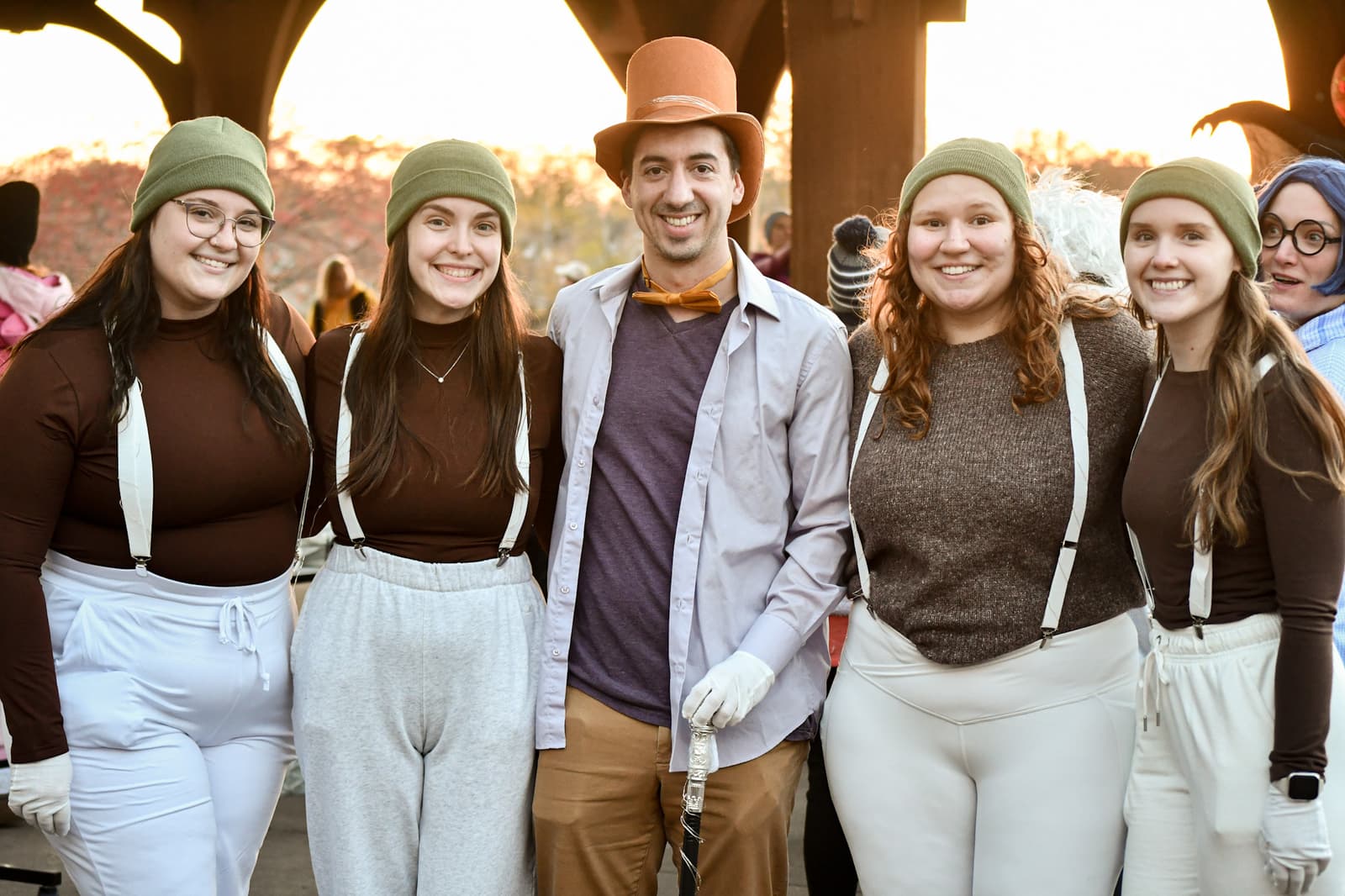 Five people in costume pose and smile for a photo. Four wear green caps, brown shirts, and white suspenders, while the person in the center wears a brown top hat, bow tie, gloves, and carries a cane. Others are visible in the background.