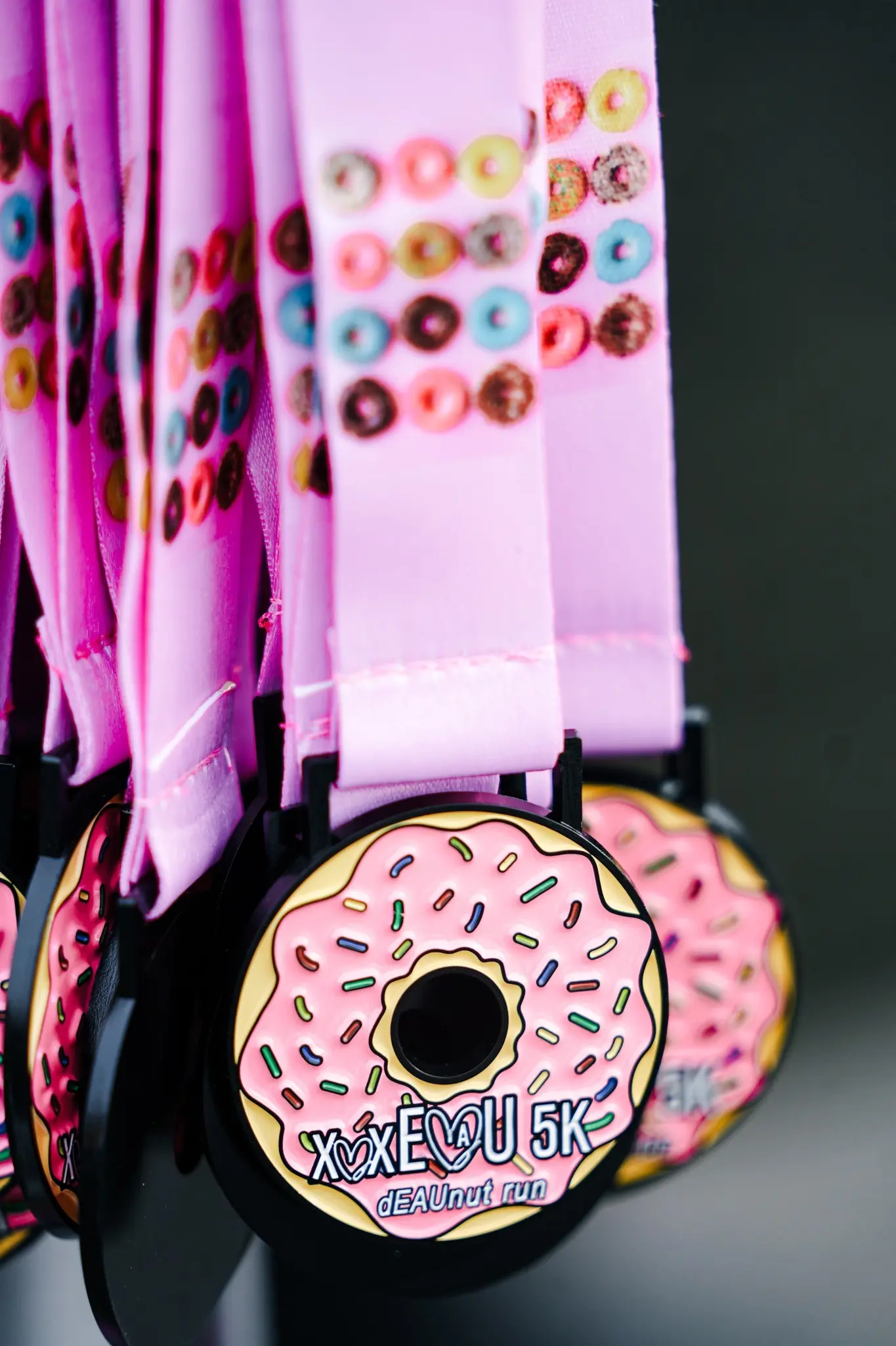 A close-up of several pink medals featuring a donut design with colorful sprinkles and "XoXo 5K DEANut run" text, hanging from pink lanyards decorated with tiny donut images.